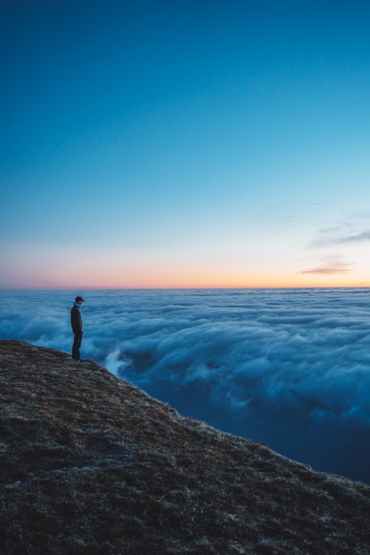Person standing on a cliff above clouds at sunset.