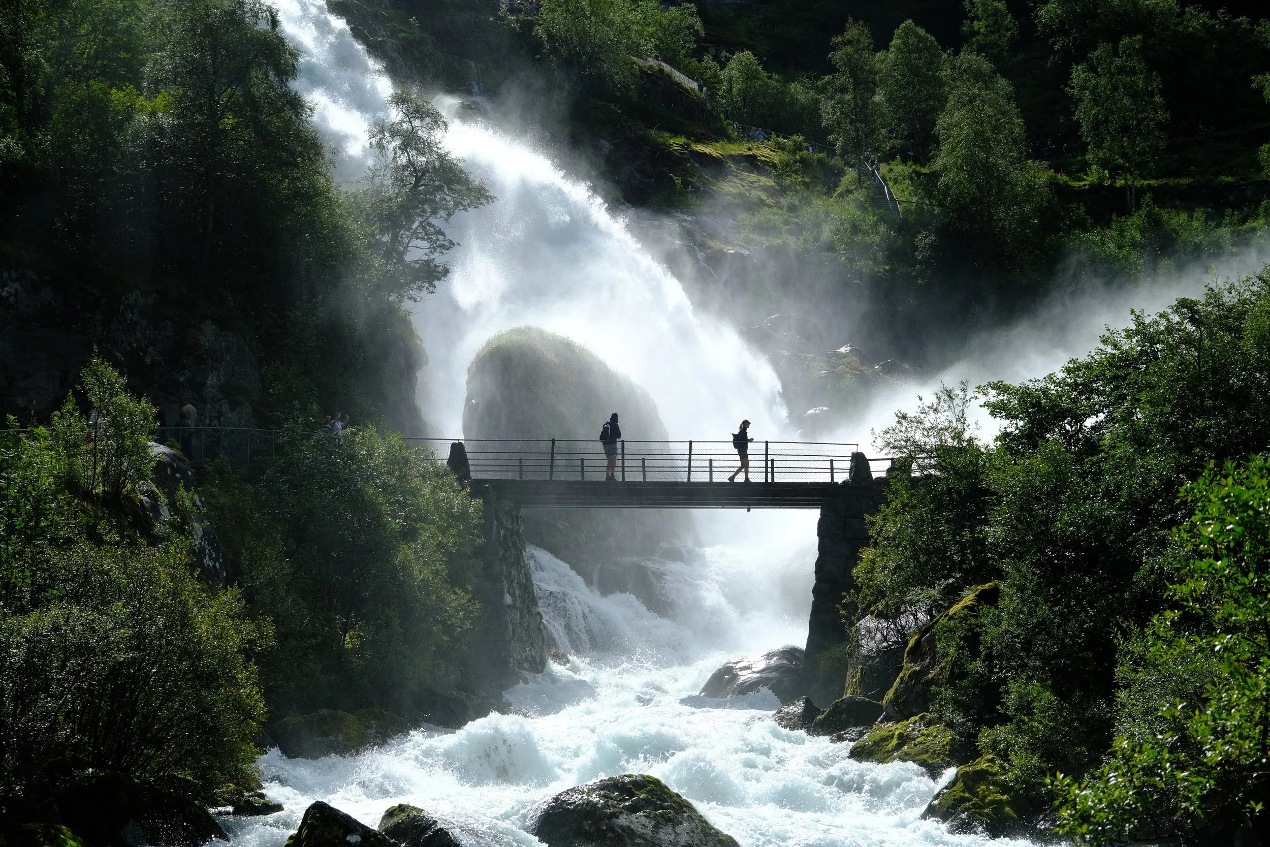 People on a bridge over a waterfall in a lush forest.