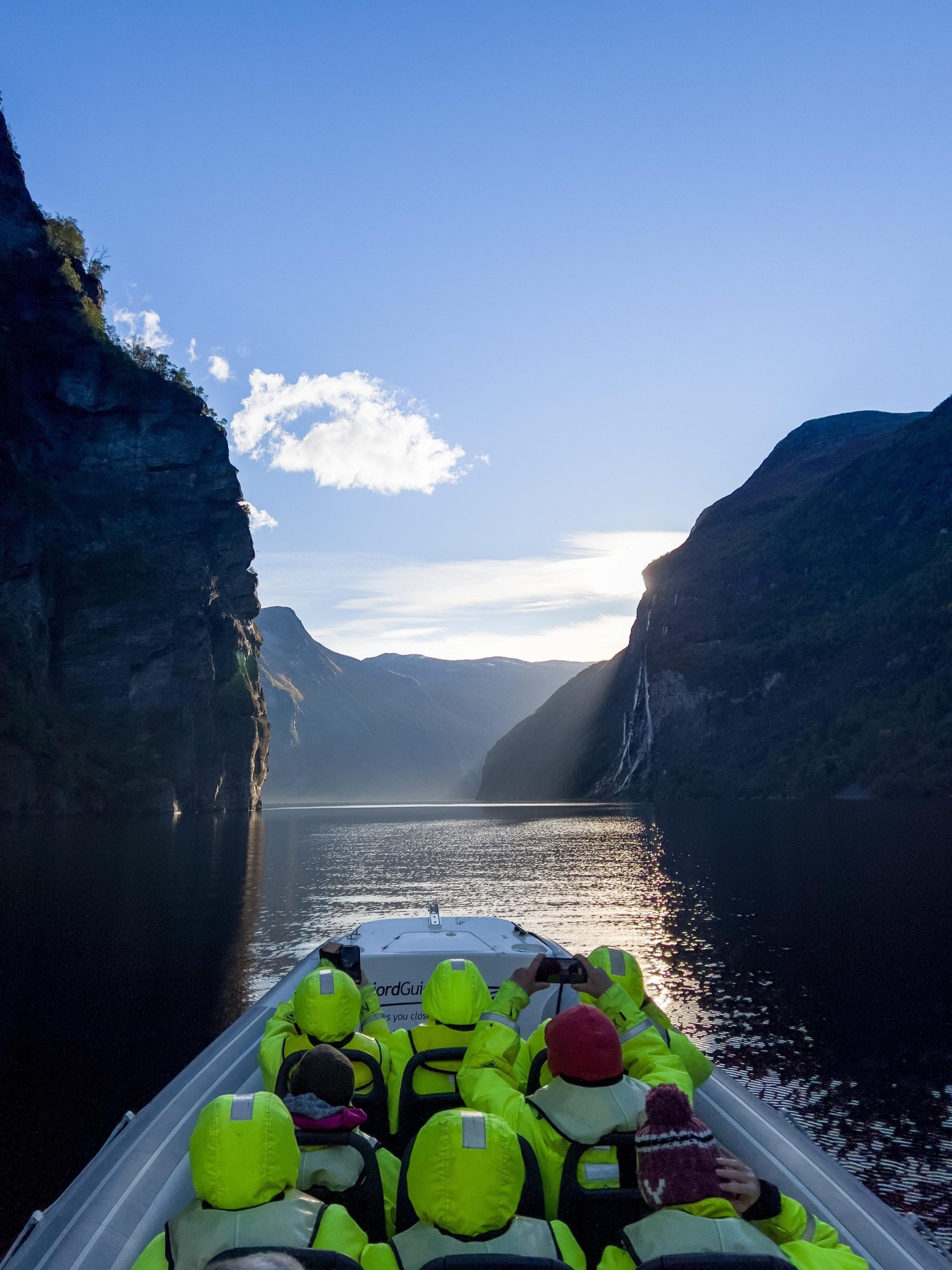 Group on boat in fjord with cliffs, waterfall, and clear sky.