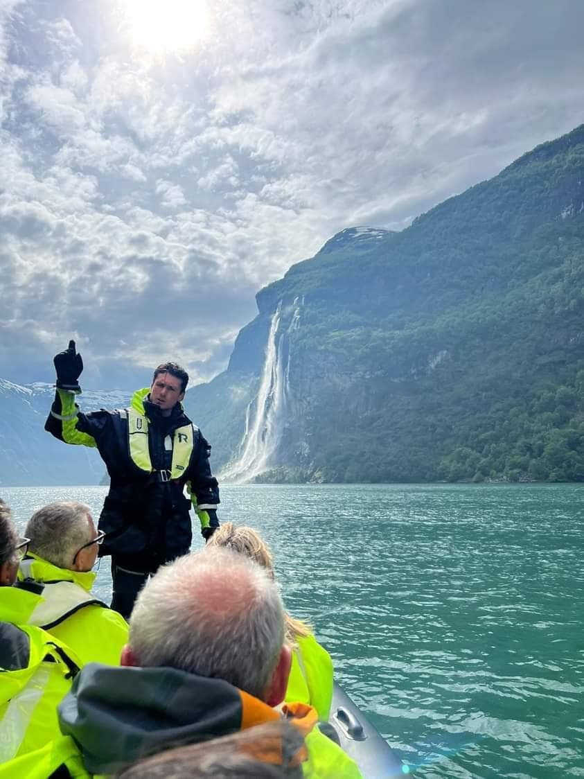 Person in a boat pointing towards a waterfall against a mountainous backdrop.