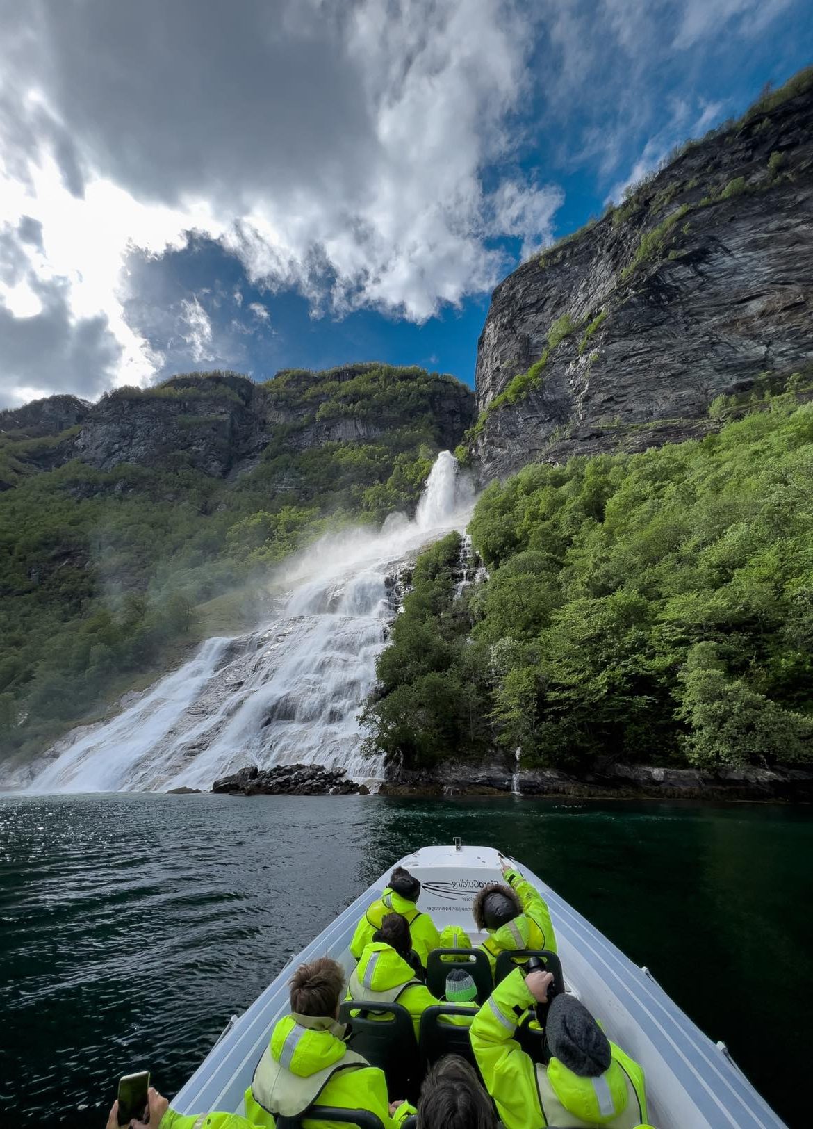 People on a boat approaching a waterfall in a mountainous landscape.