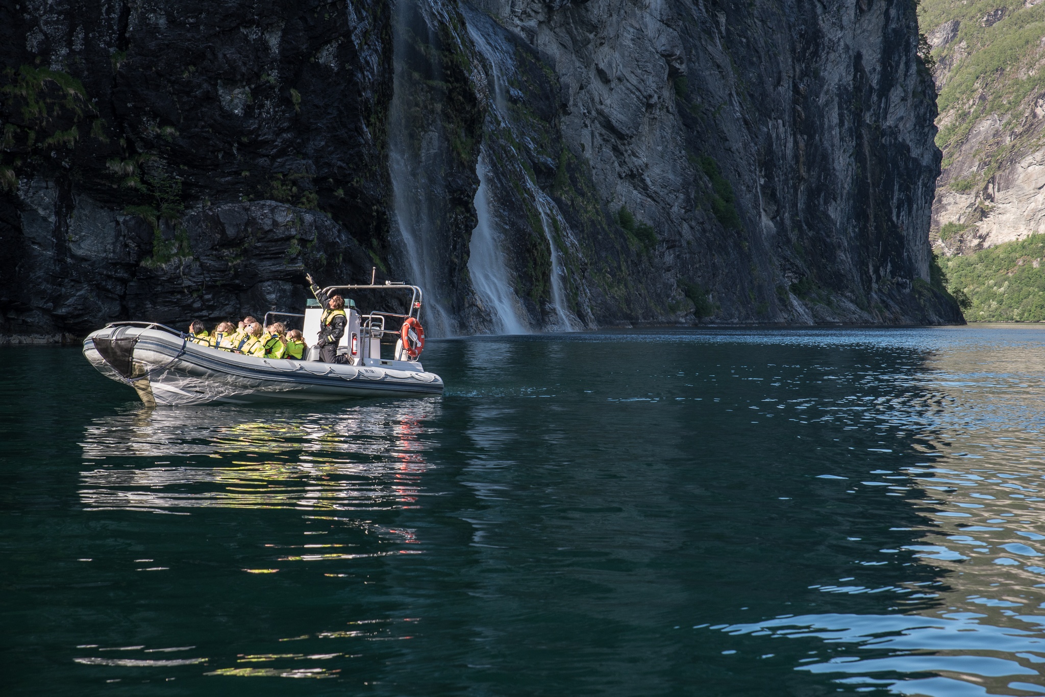 Inflatable boat with people near waterfall and cliffs on a calm body of water.