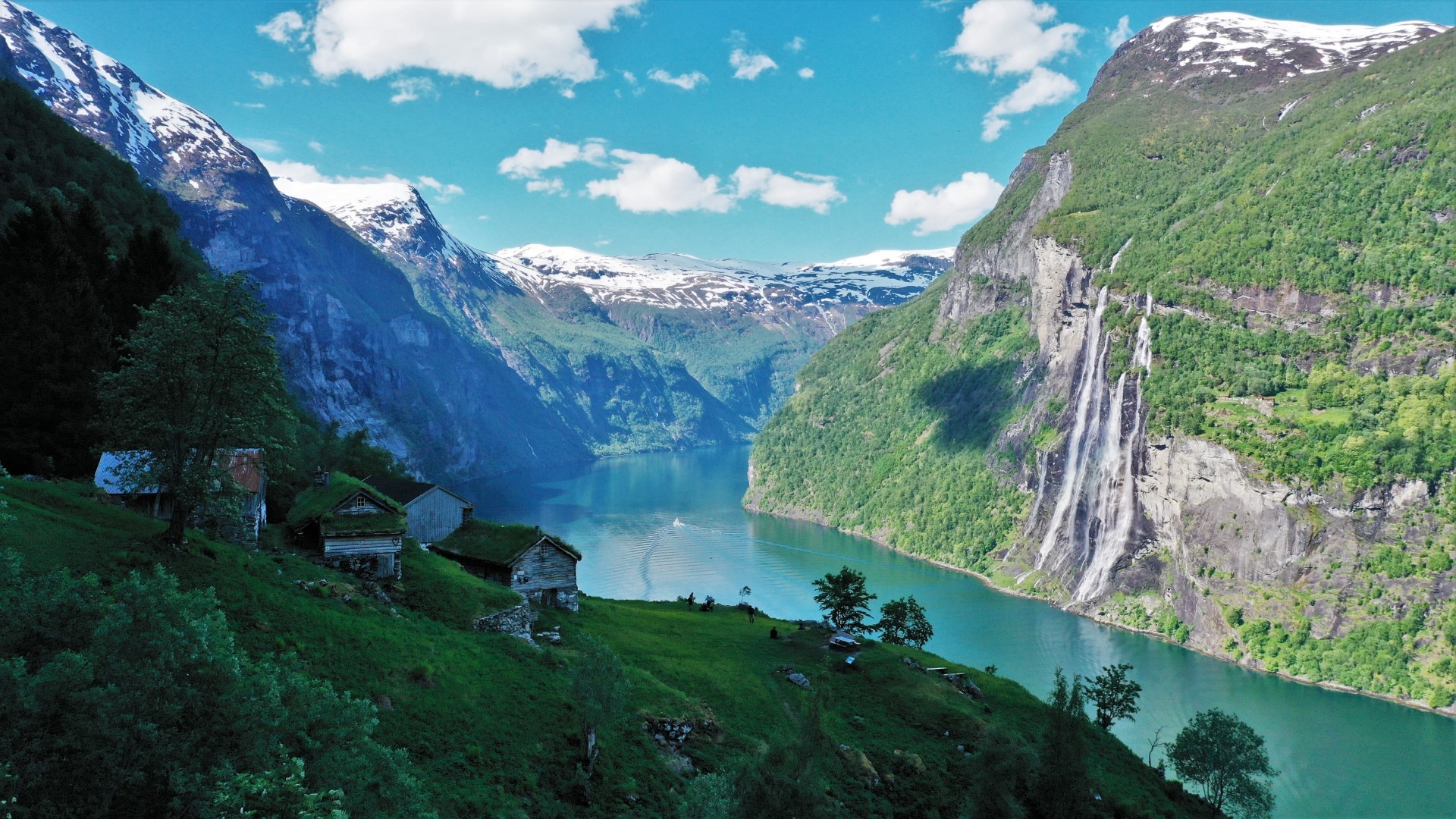 Scenic fjord with steep cliffs, waterfall, and small houses on grassy slope.