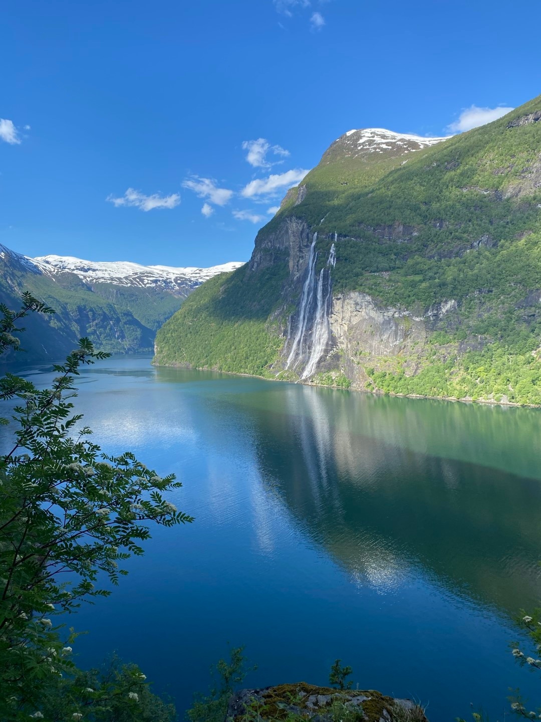 Serene fjord with waterfall, lush greenery, and mountain backdrop under a clear blue sky.