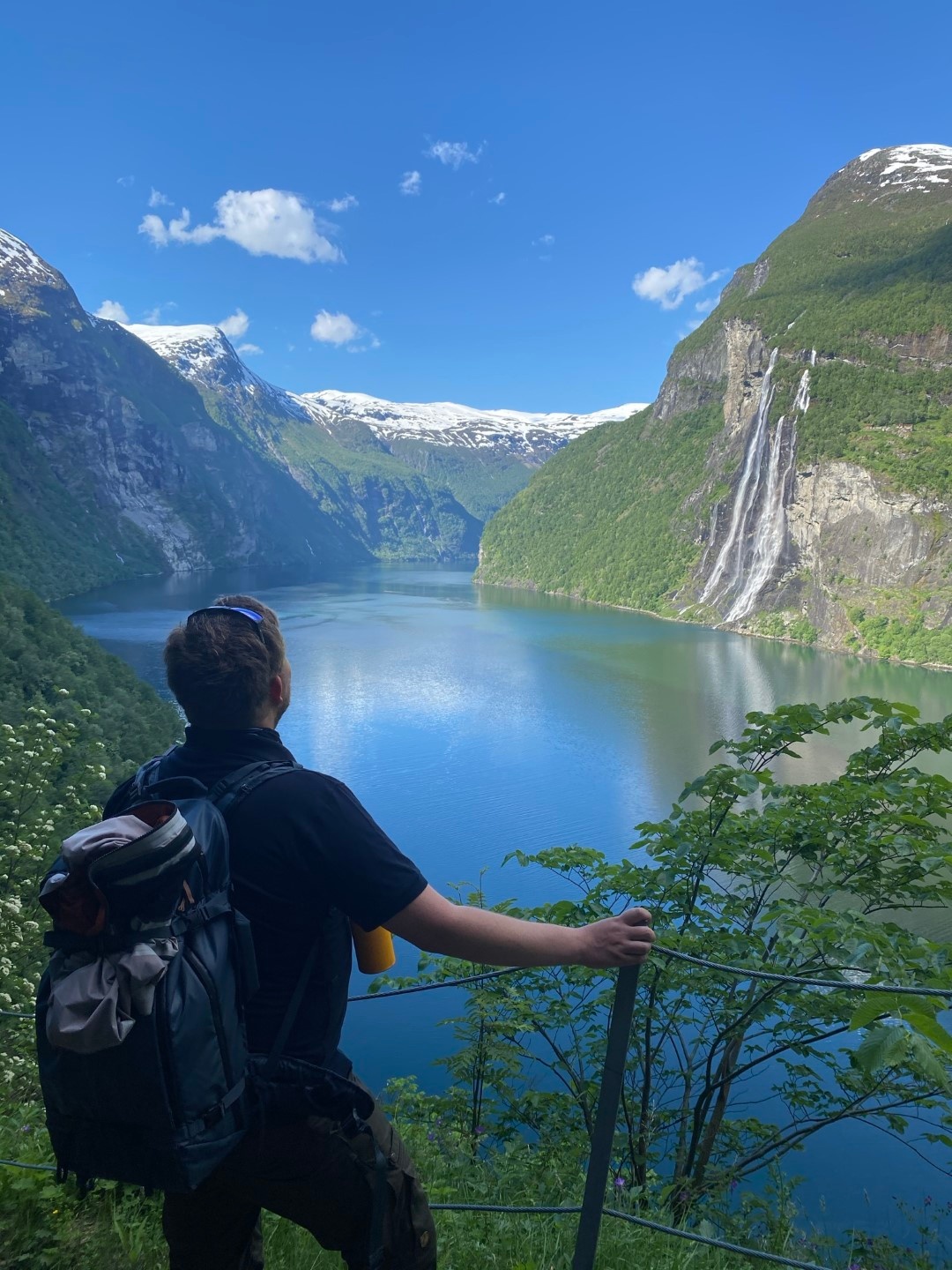 Person with backpack admiring scenic fjord view with waterfall and mountains.