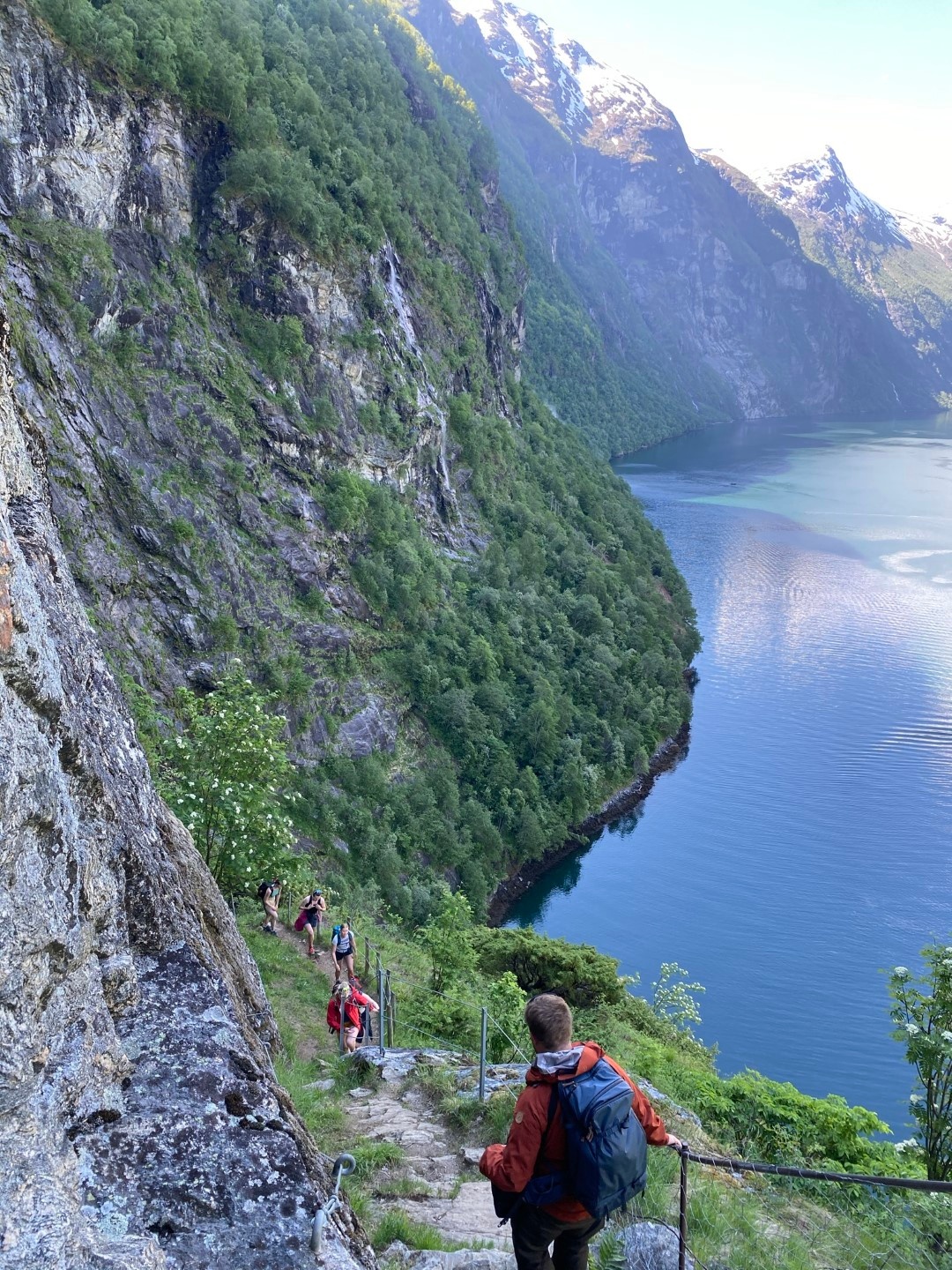 Hikers walking on a narrow cliffside trail above a lake in a mountainous landscape.