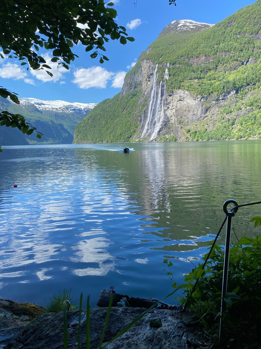 Scenic fjord with waterfall and mountains under a blue sky, seen from a shaded area with foliage.