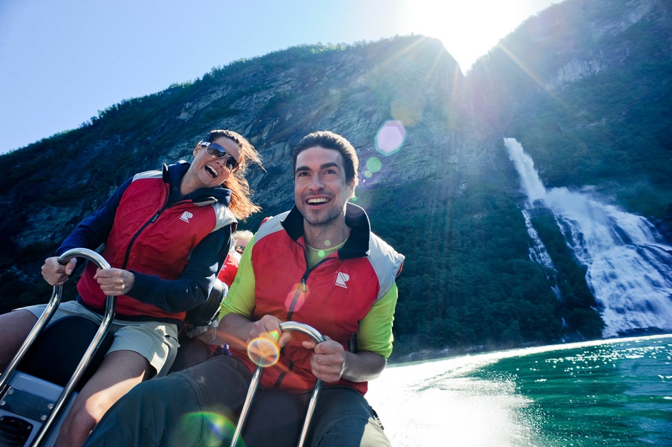 Two people in red vests enjoying a boat ride by a waterfall on a sunny day.
