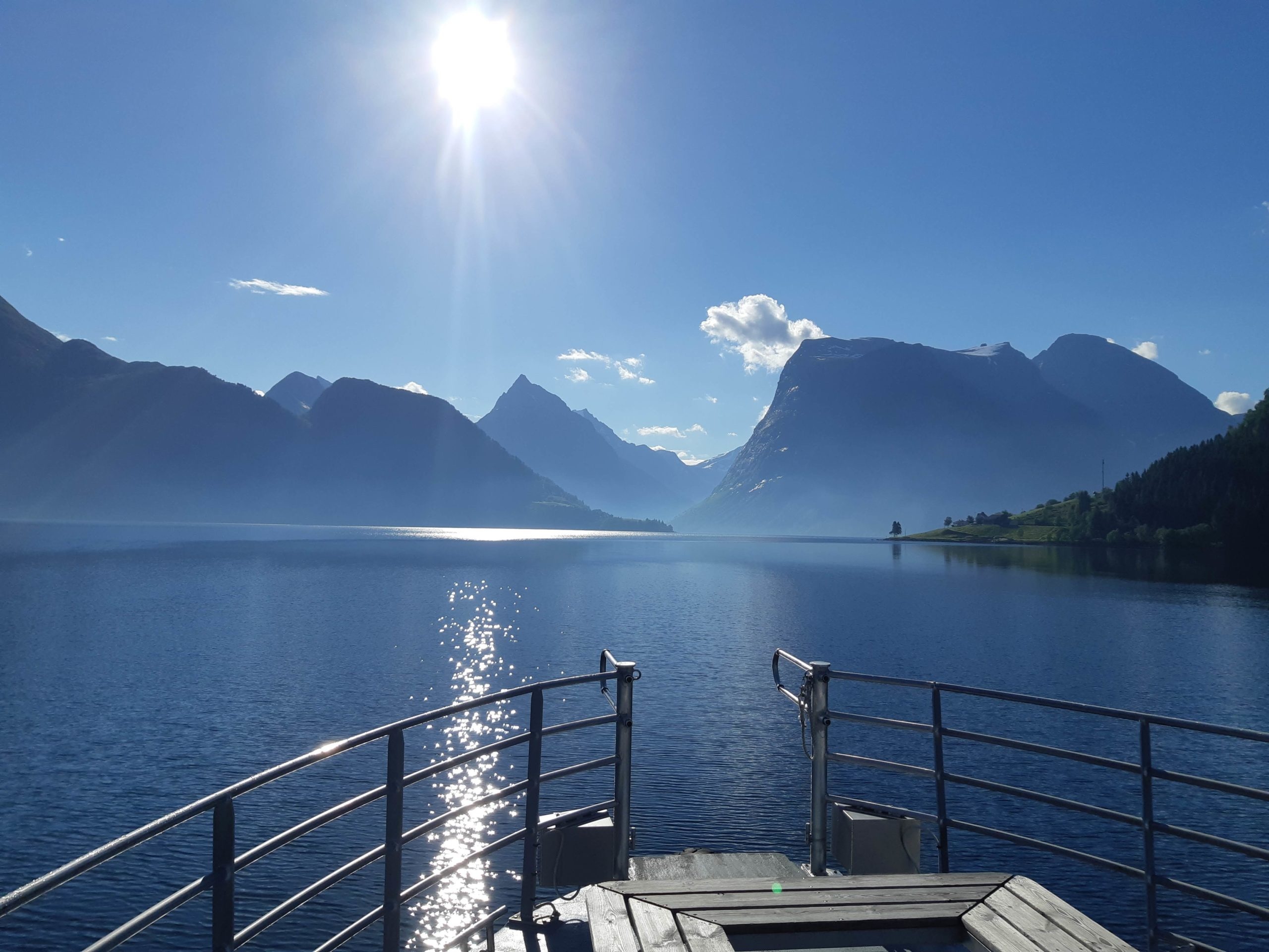 Sunny lake view with mountains and a boat railing in the foreground.