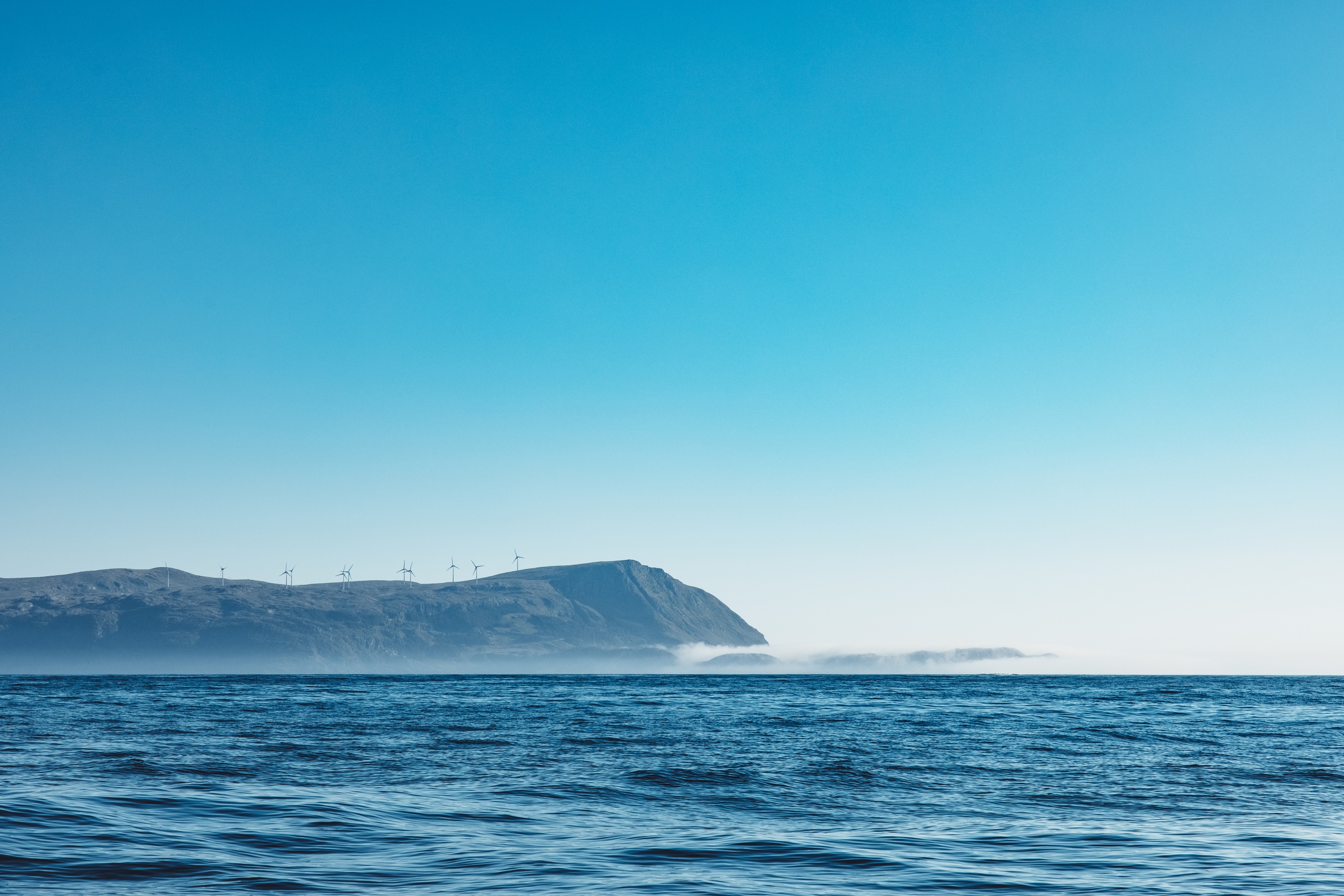Ocean view with distant hills and wind turbines under a clear blue sky.