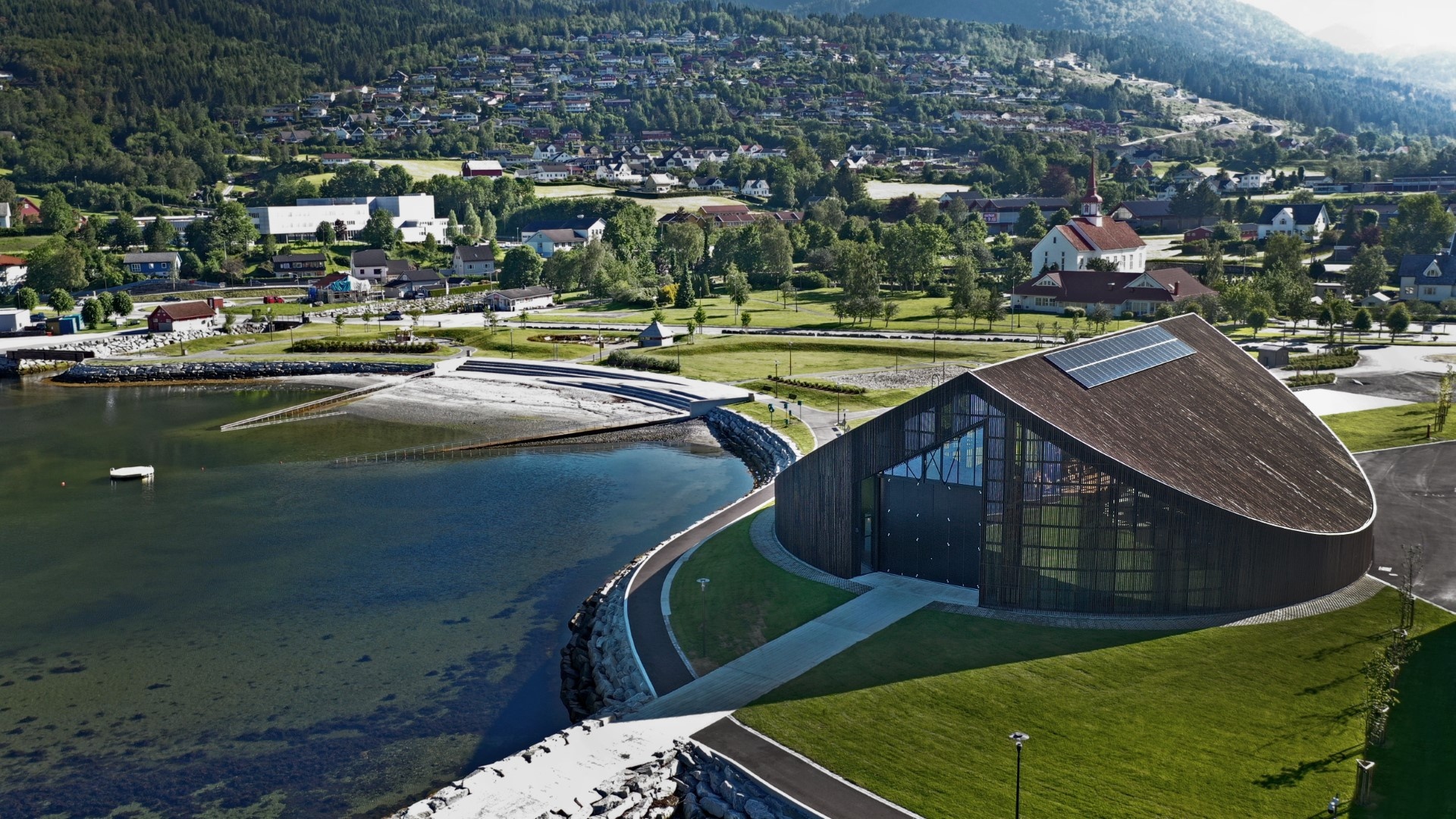 Modern building by a lake with green hillside and town in background.