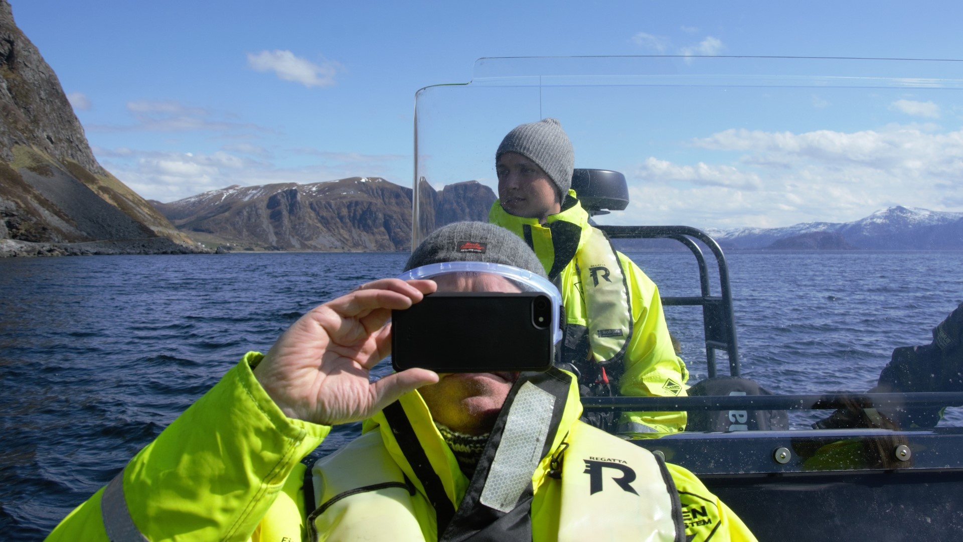Two people in yellow jackets on a boat; one takes a photo, mountains in the background.