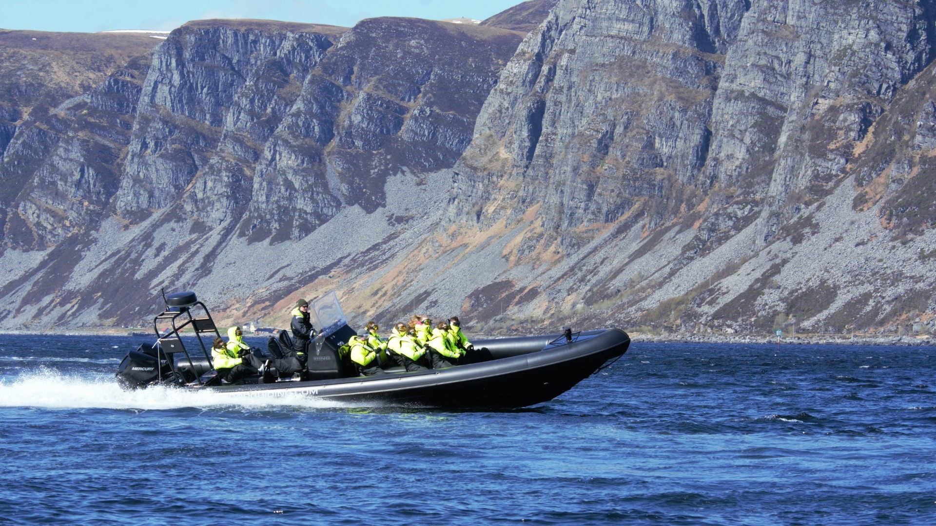 Tourists in bright jackets on a speedboat near rocky cliffs.