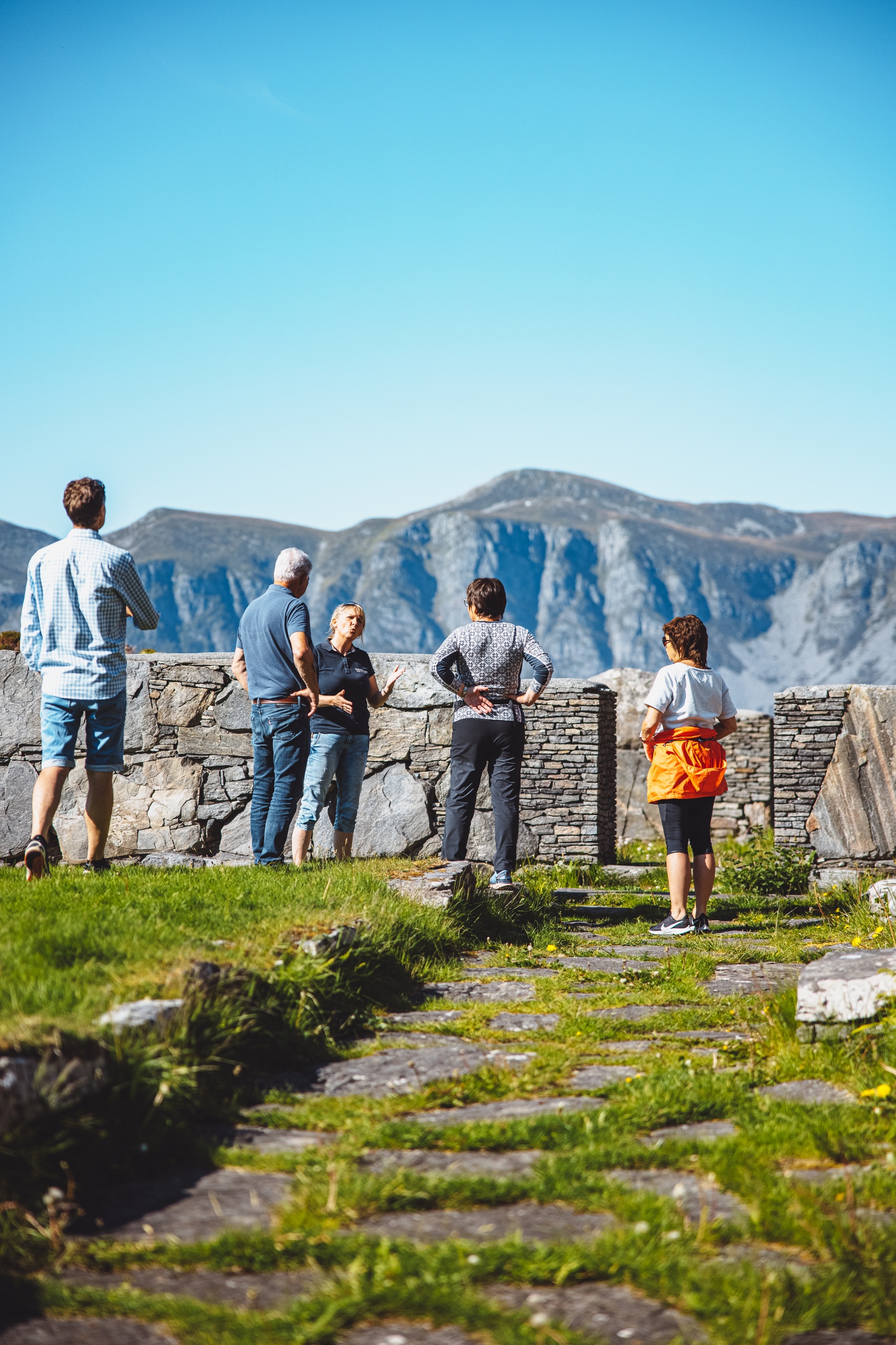 People standing on a stone path near a stone wall, with mountains in the background.