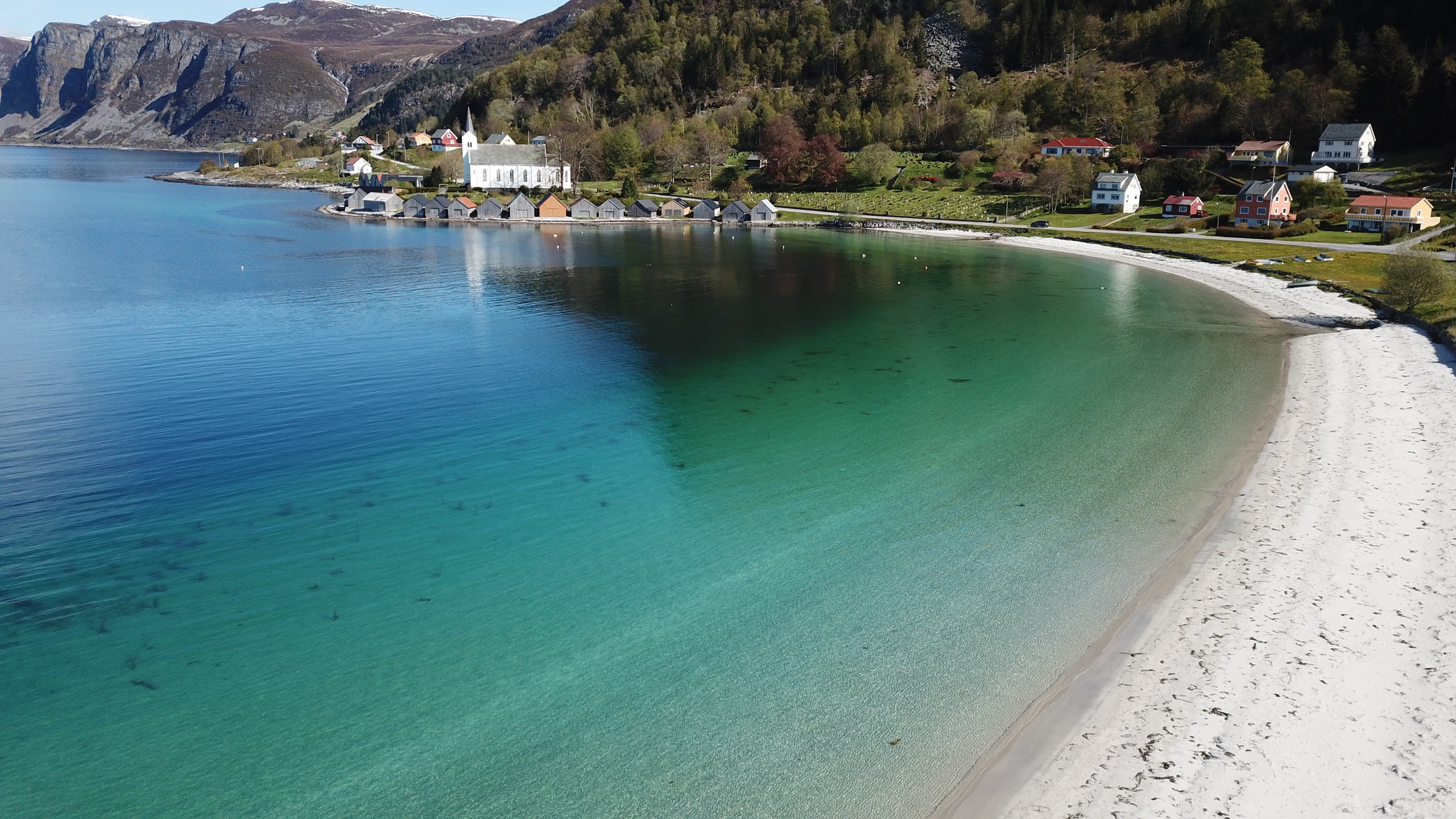 Coastal village with white church, colorful houses near a clear blue sea and sandy beach.