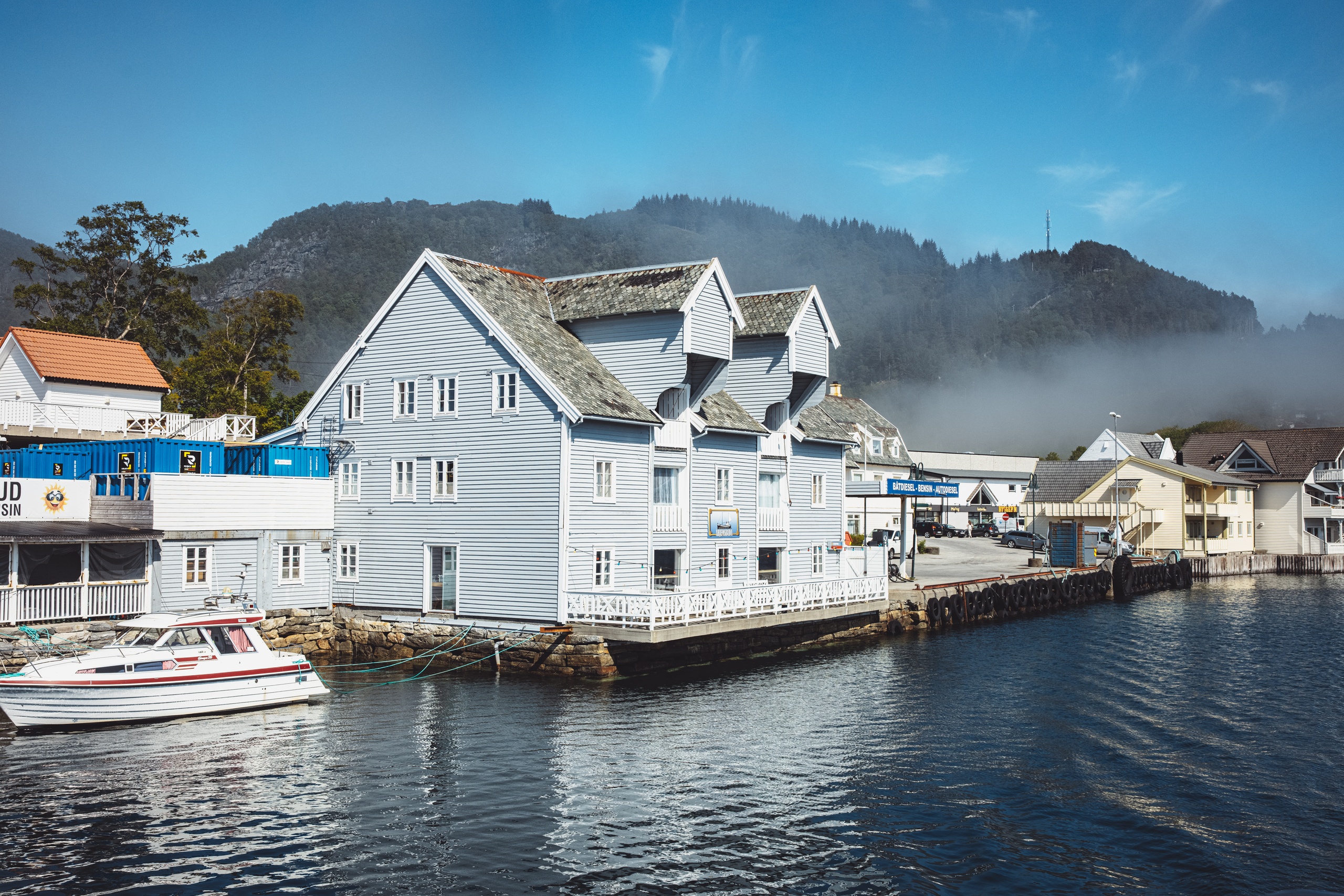 Wooden buildings by a calm river with foggy mountains in the background.