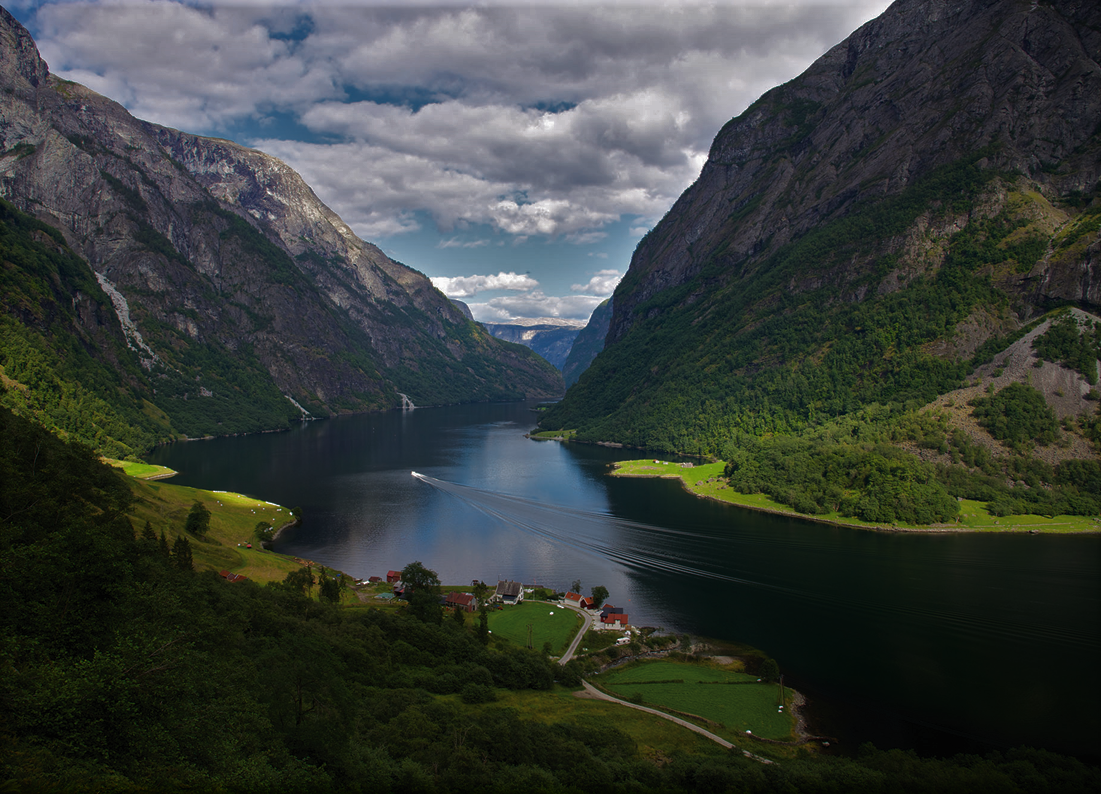 Scenic fjord with steep mountains, a boat, and a small village on the shore under a cloudy sky.