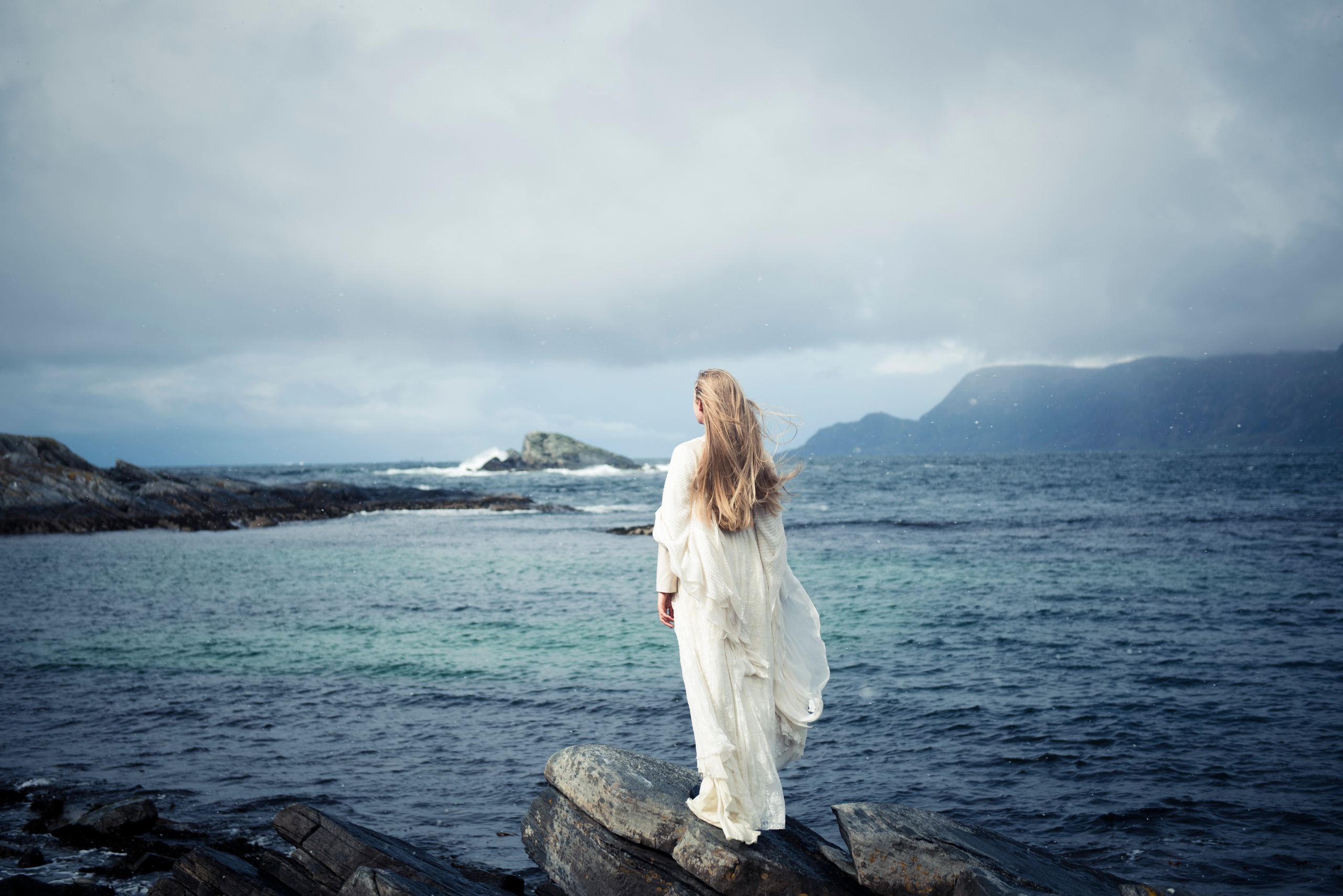 Woman in white dress stands on rocky shore, looking at the sea.