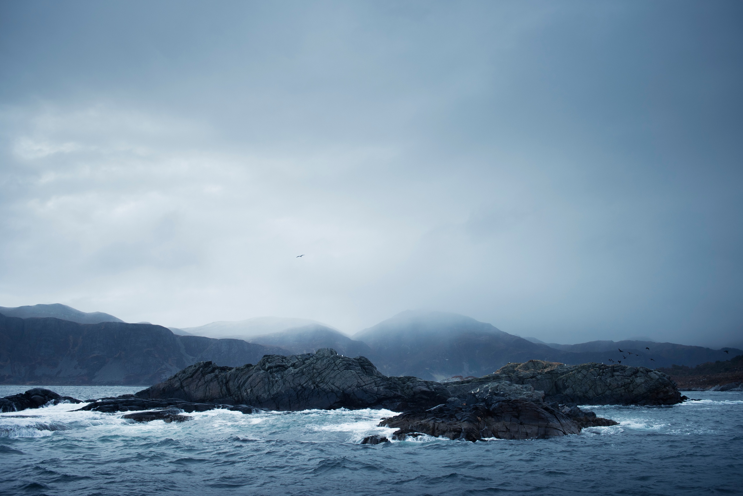Rocky coastline with waves under cloudy sky and distant mountains.