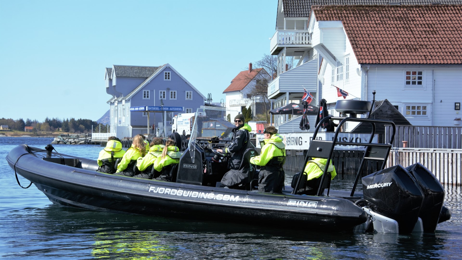People in yellow jackets on a black boat near dockside houses.