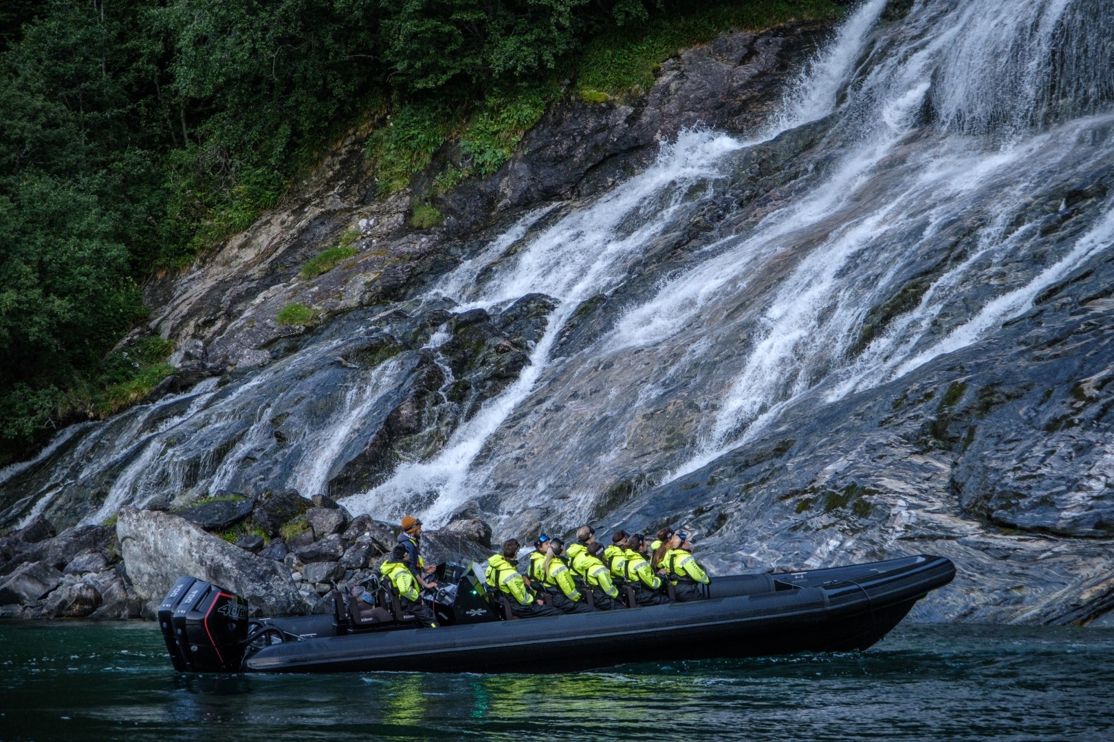 Group in yellow jackets on a black boat near a rocky waterfall.