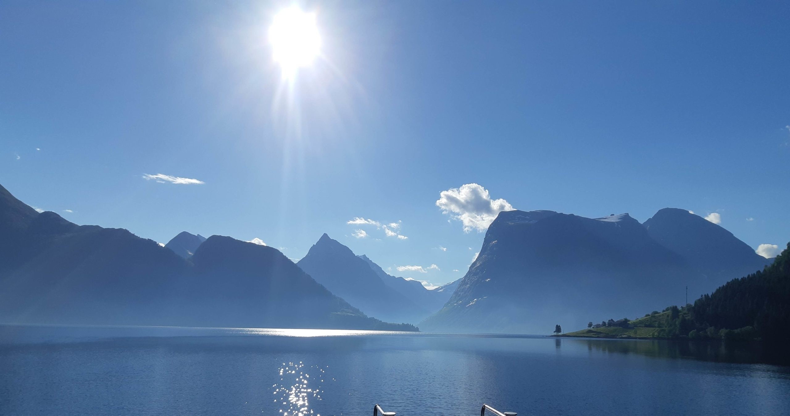 Scenic lake view with bright sun above mountains and clear blue sky.