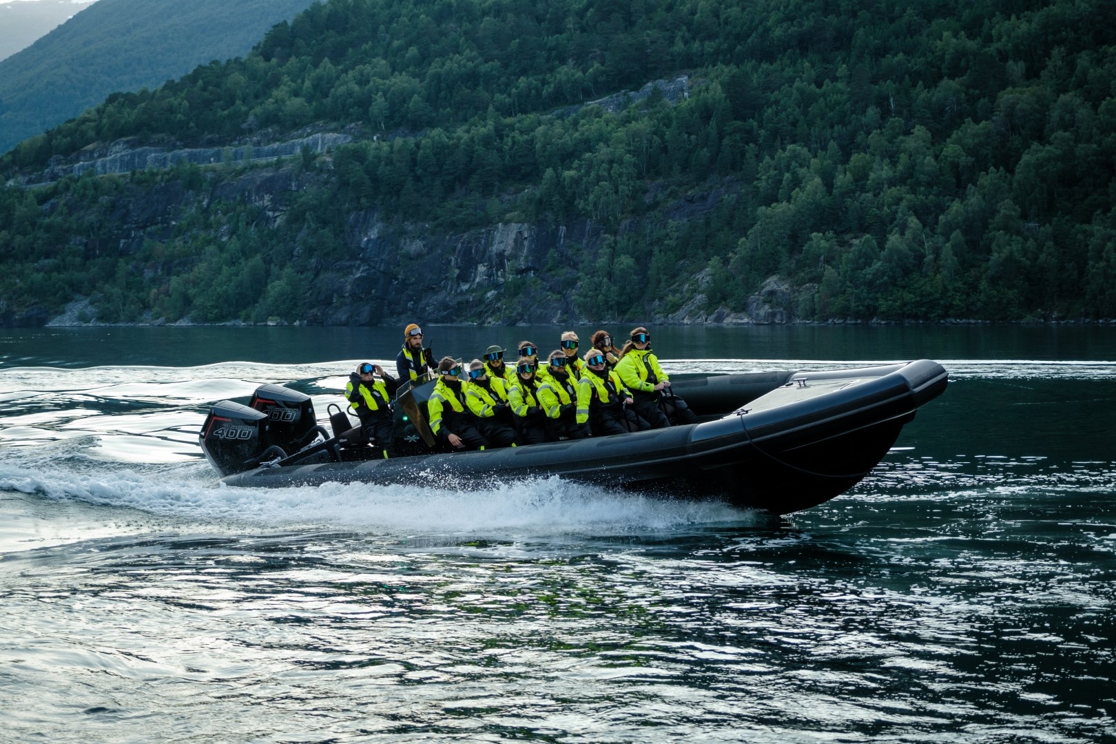 Group on fast black boat in a fjord, wearing yellow jackets, with forested mountains in background.