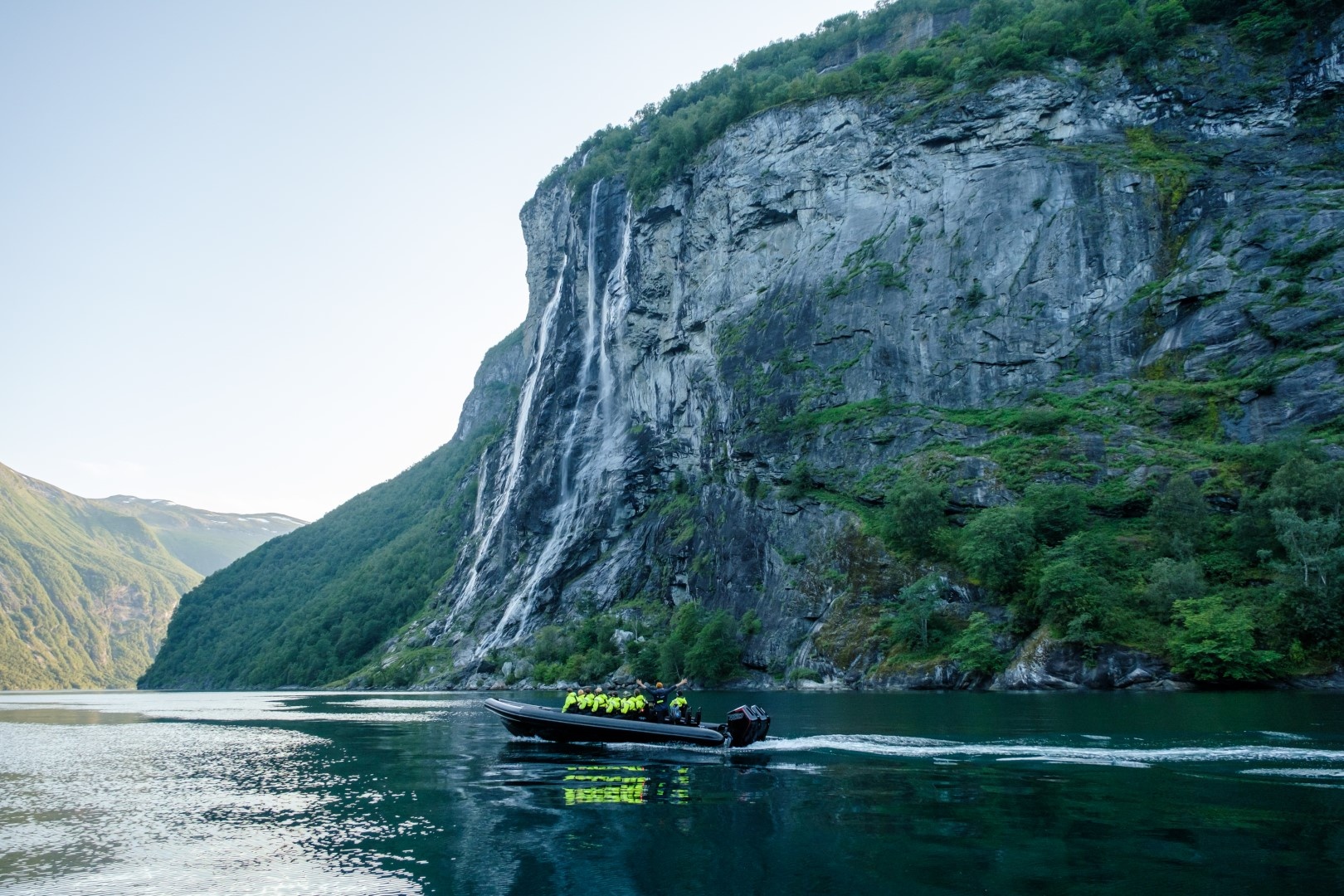 Boat with people near a waterfall in a fjord under clear sky.