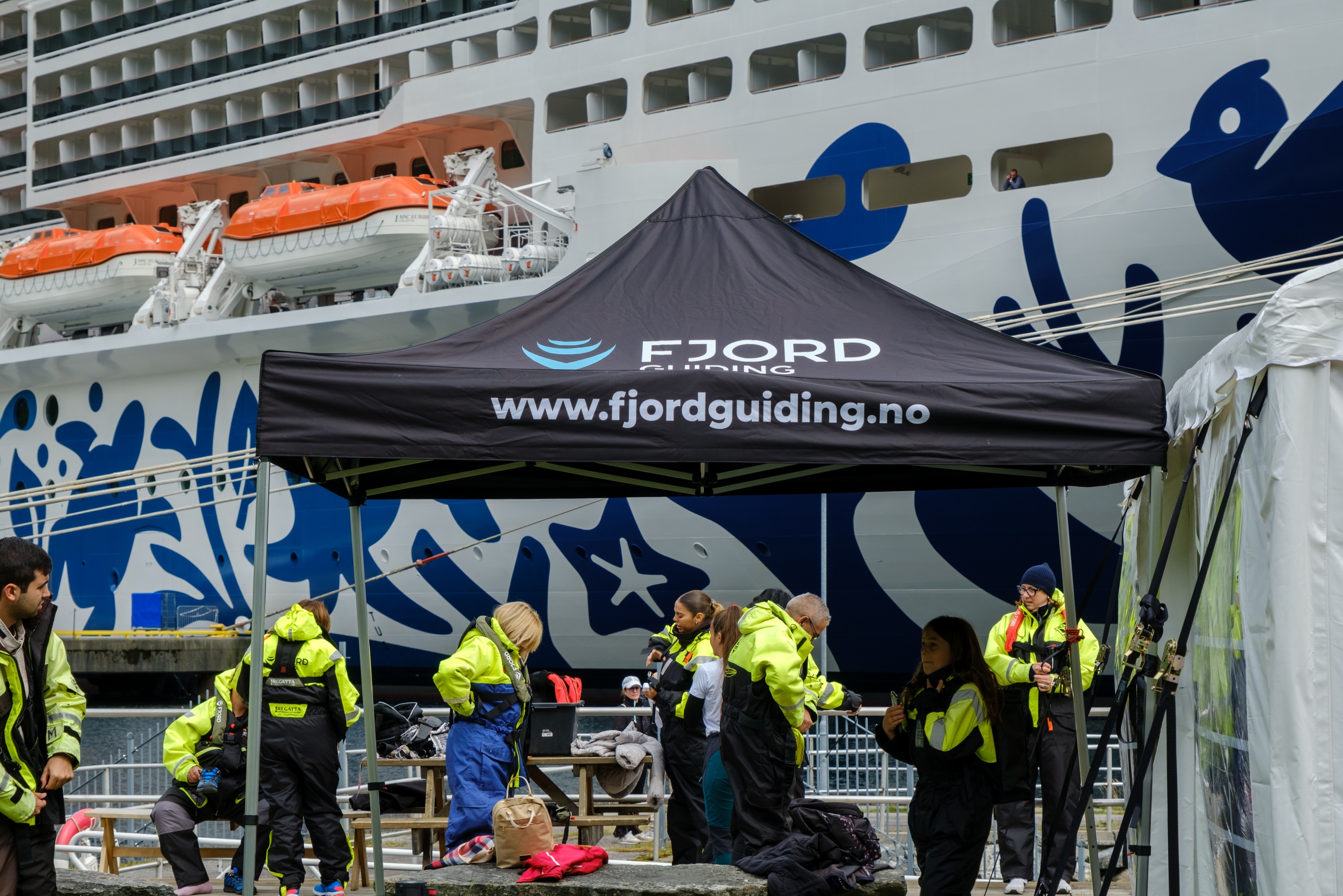 People in yellow jackets under a tent with fjord tour branding near a cruise ship.
