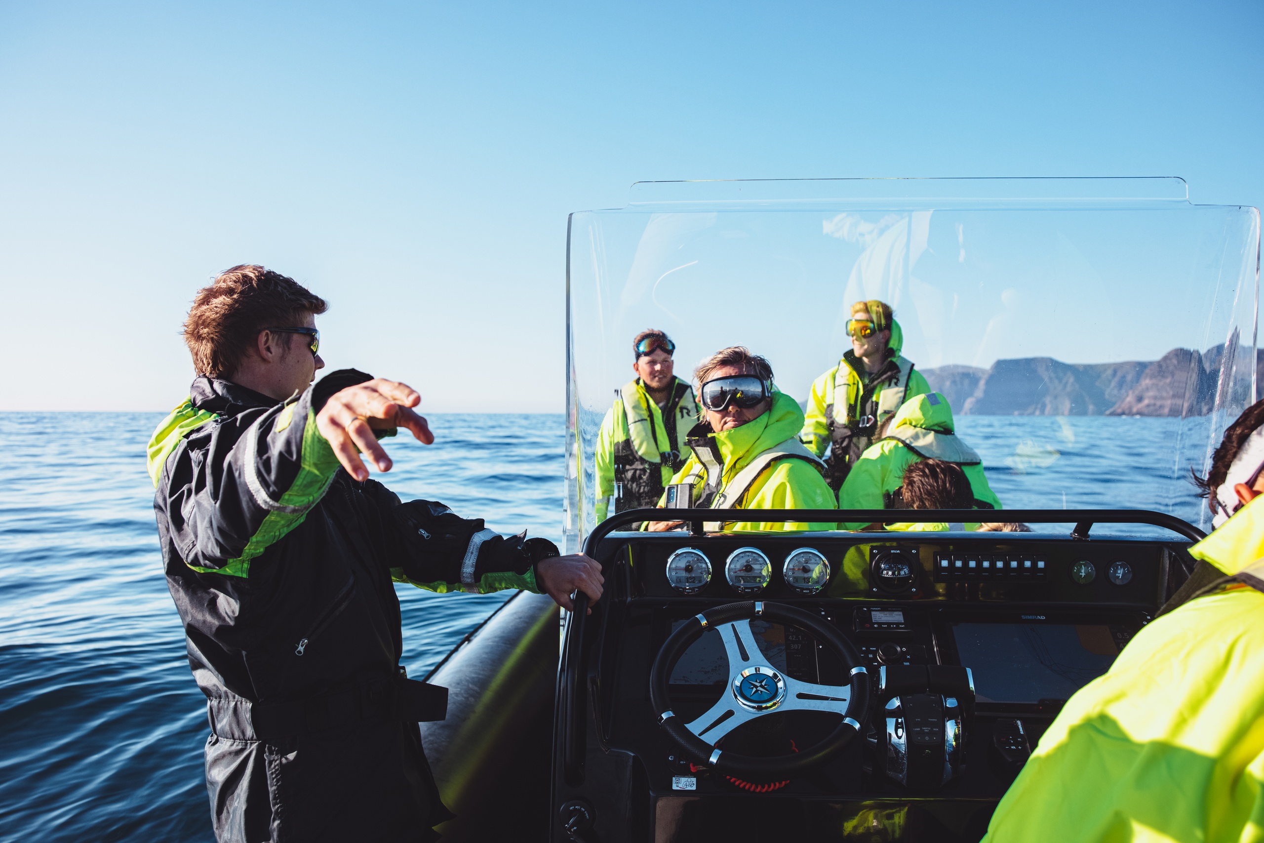People in bright jackets on a boat in calm ocean waters with mountainous backdrop.
