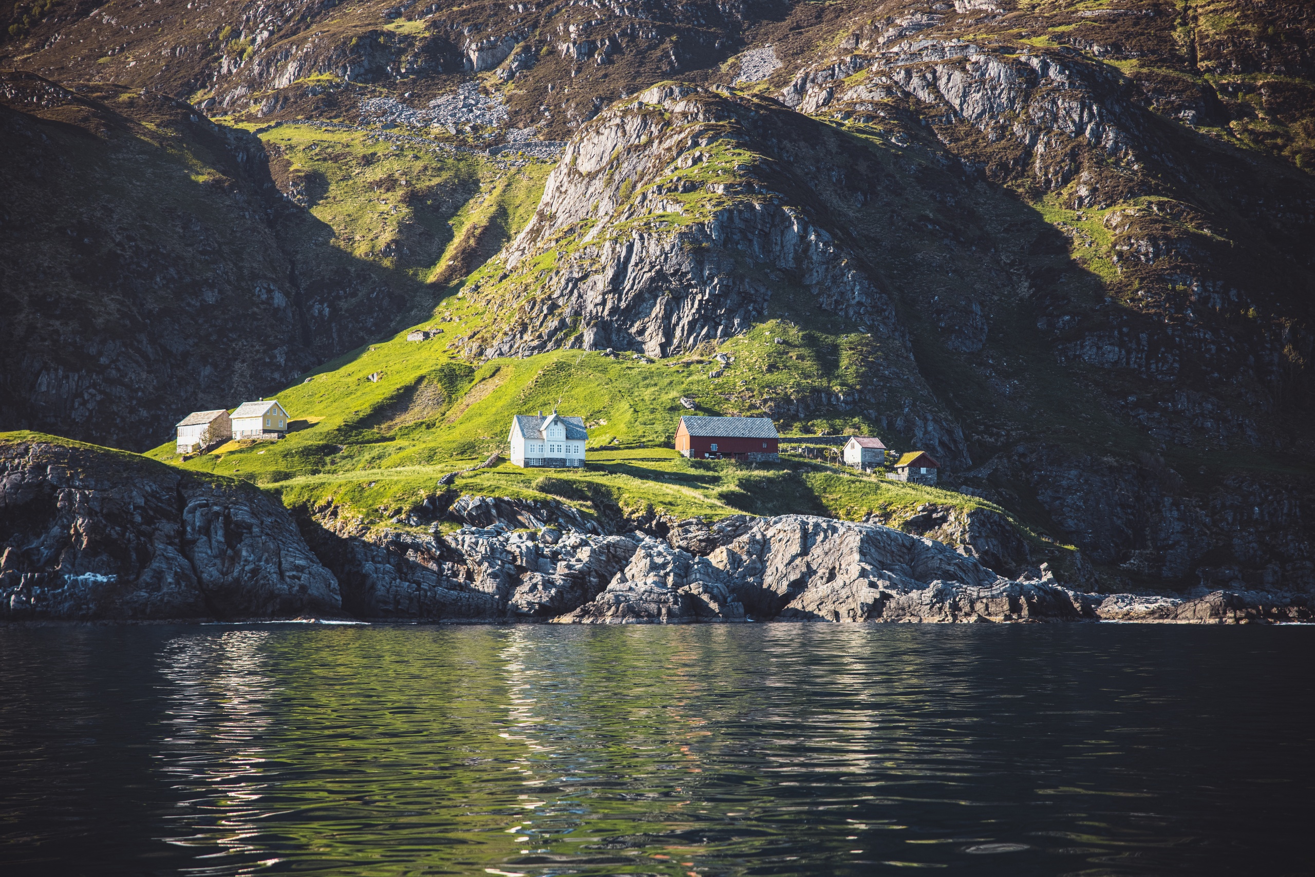 Houses and green hills by rocky coastline with calm water.