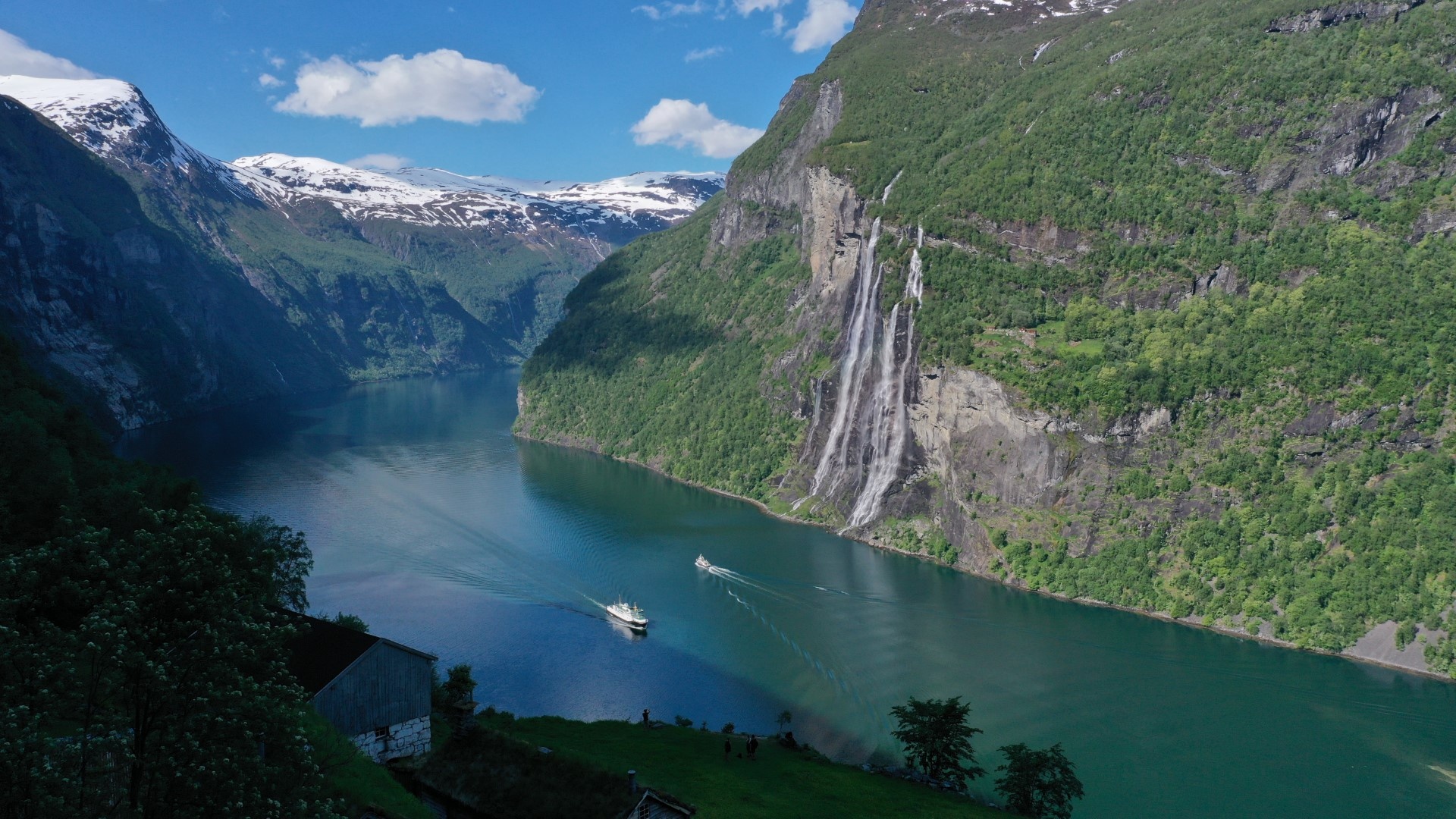 Scenic fjord with boats, waterfall, green hills, and snow-topped mountains.