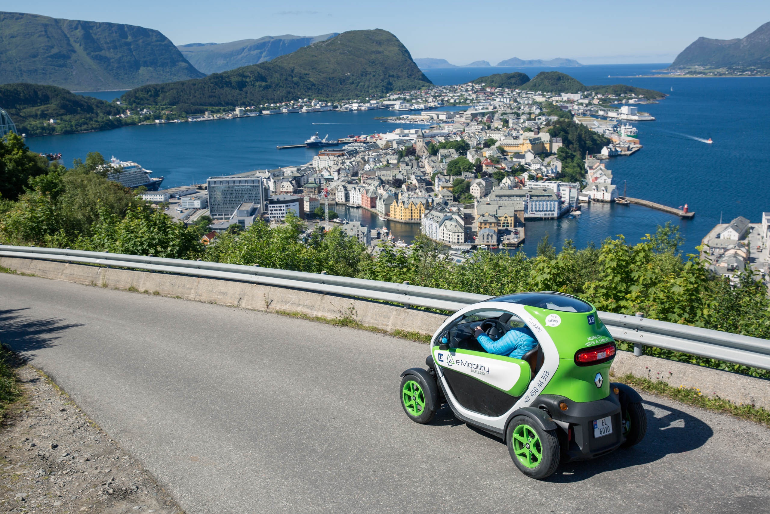 Small green car on a scenic mountain road overlooking a coastal town and fjord.