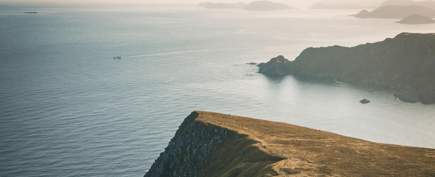 Coastal cliff landscape with sea and distant islands under a hazy sky.