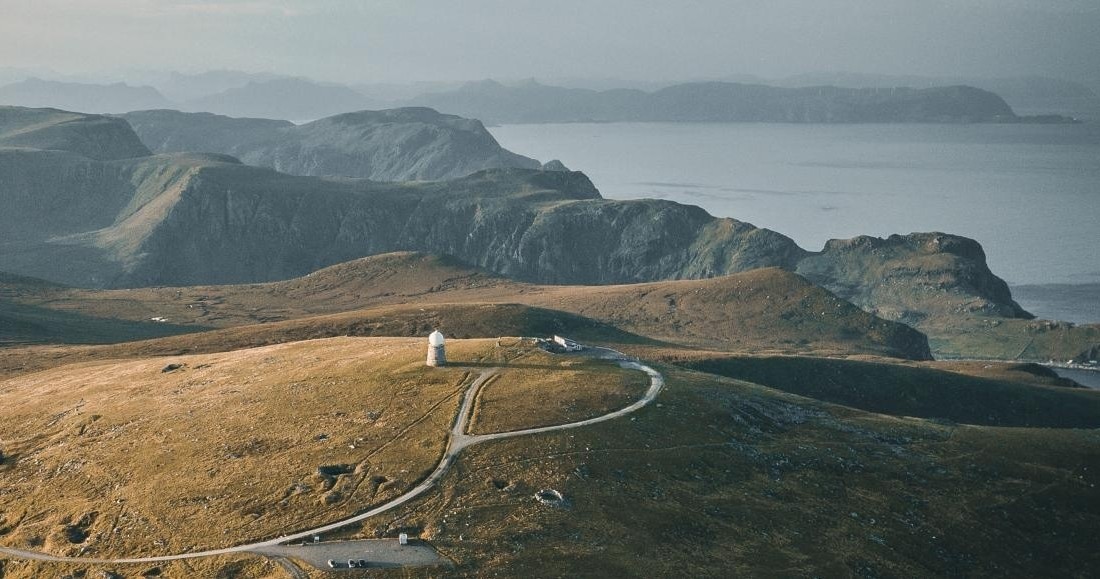Coastal landscape with hills, winding road, and small lighthouse.