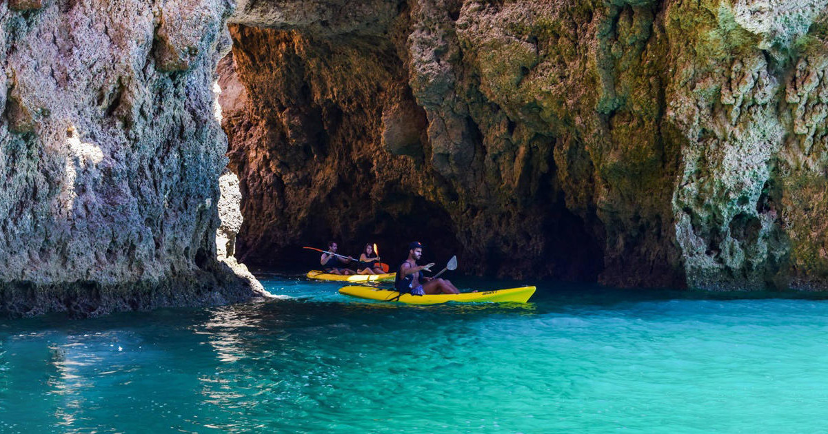 Kayakers paddling through a rocky sea cave on clear turquoise water.
