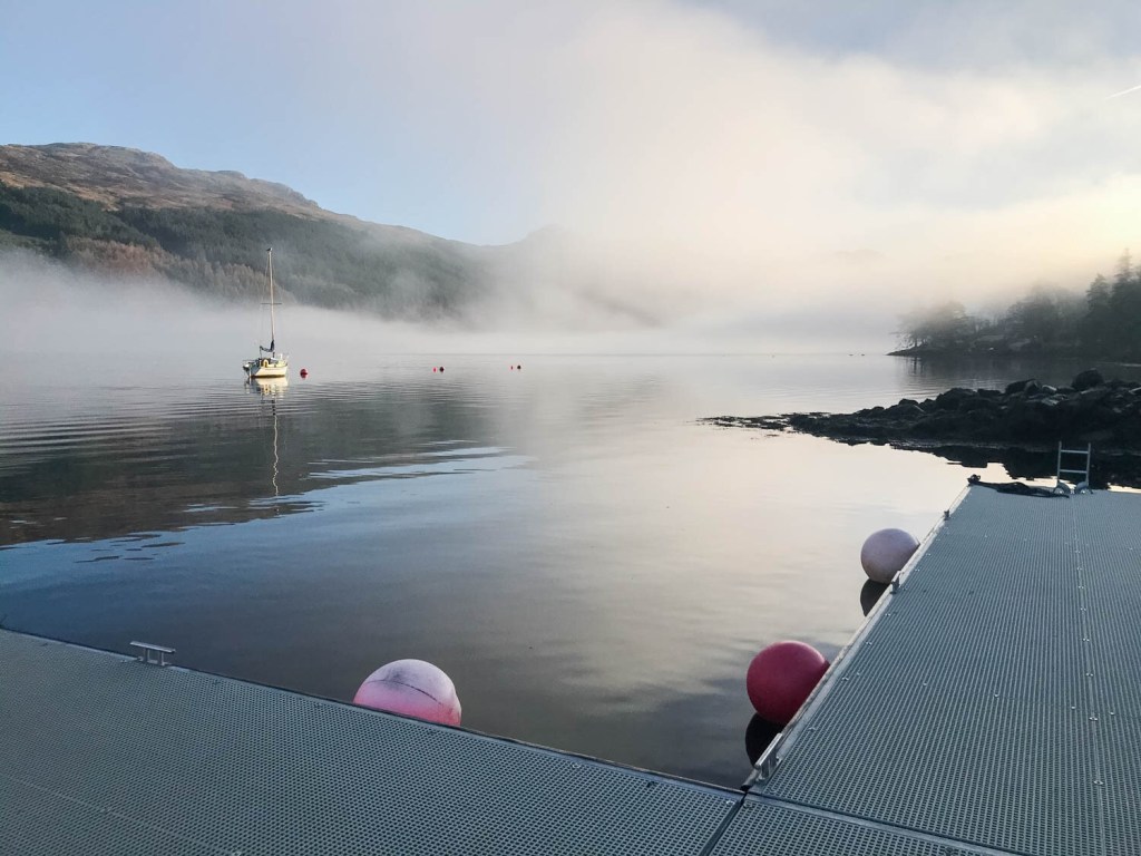 Misty lake with boat, dock, buoys, and mountainous background.