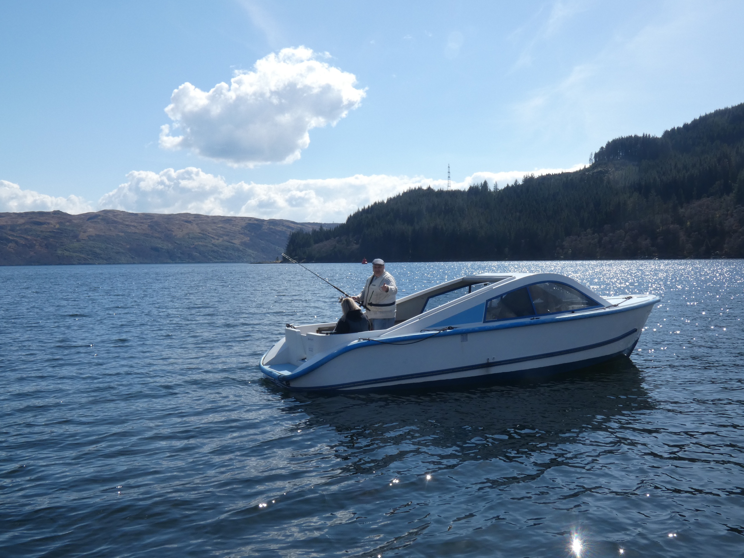 Person fishing on a small white boat in a lake with hills in the background.