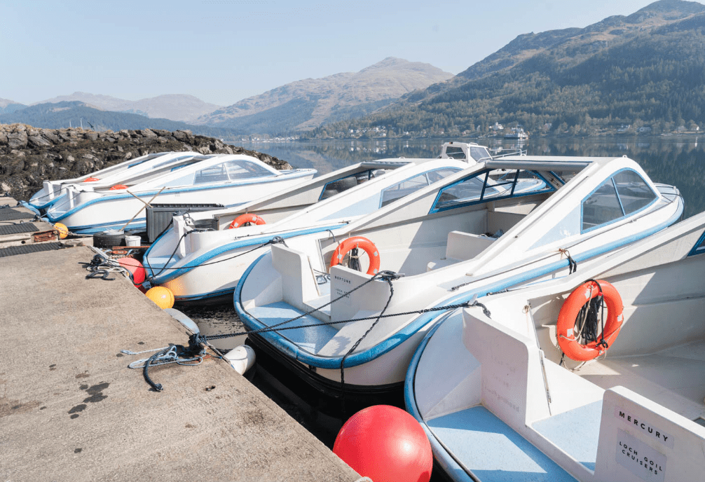 Four white boats docked by a mountain lake with life buoys and orange buoys.
