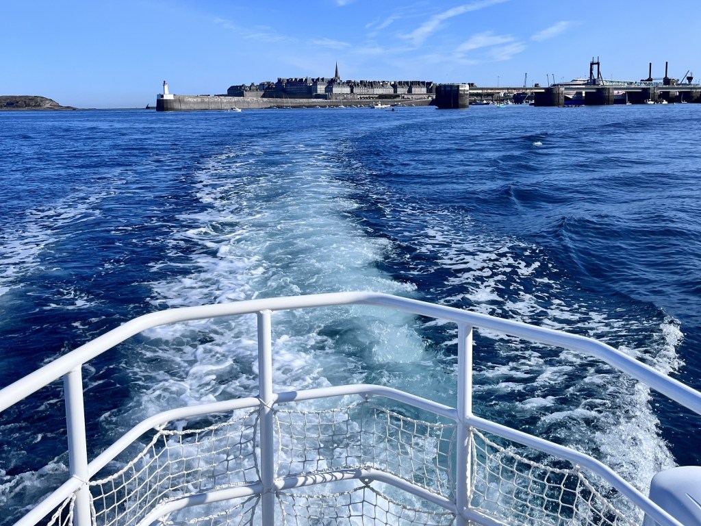 View from boat with wake in water, distant shoreline with lighthouse and buildings under blue sky.