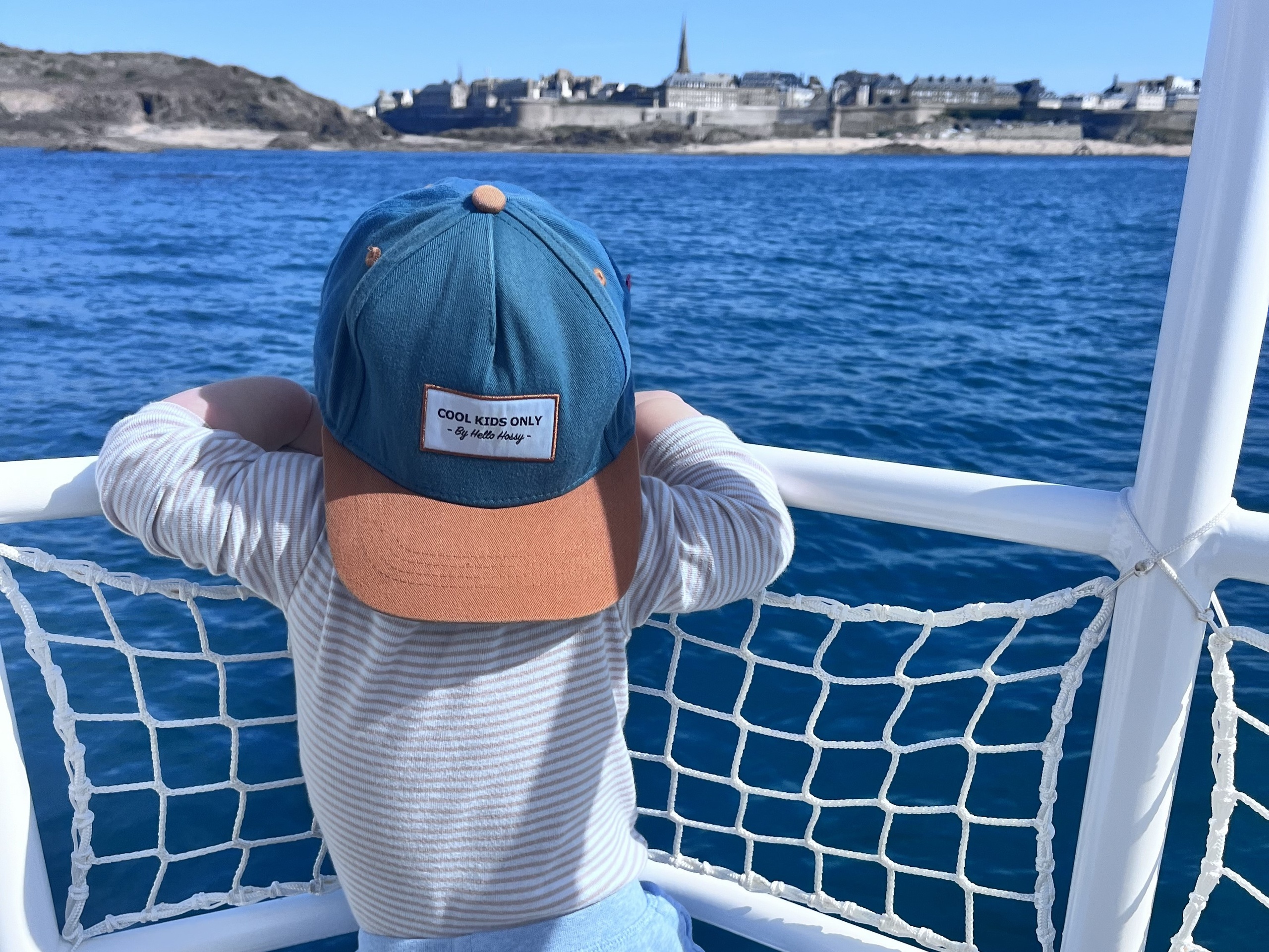 Child in a cap looking at the sea from a boat with a distant city view.