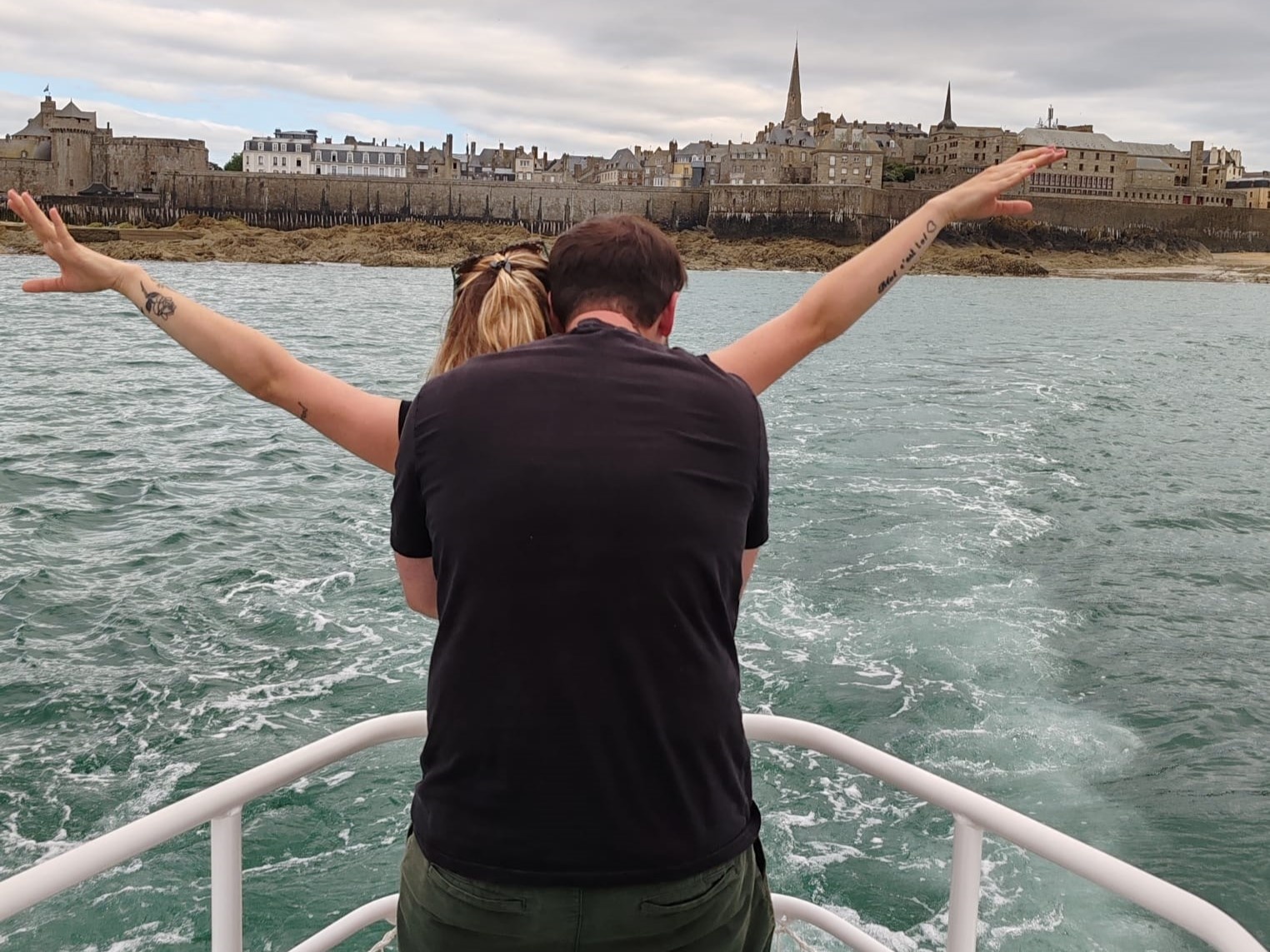 Couple on a boat recreating Titanic pose, with a coastal town in the background.