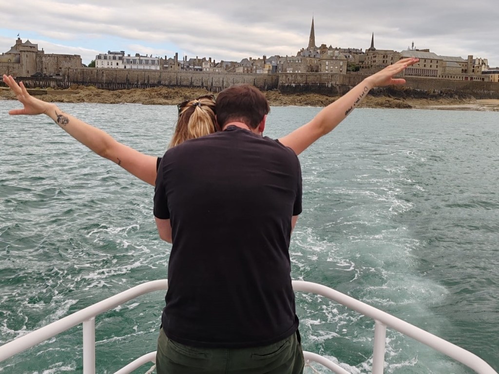 Couple on a boat recreating Titanic pose, with a coastal town in the background.