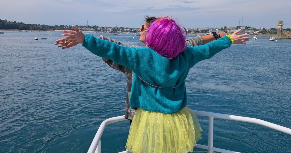 Person with pink wig and colorful clothes on a boat, arms outstretched towards the sea.