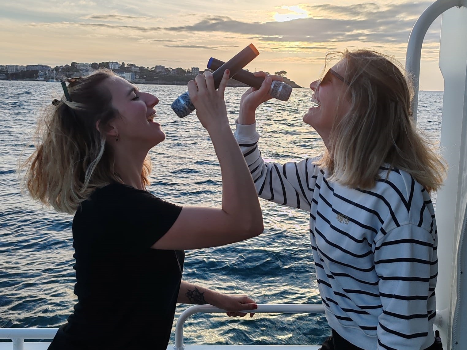 Two people smiling on a boat, holding metal items like microphones, with a sunset over the sea.