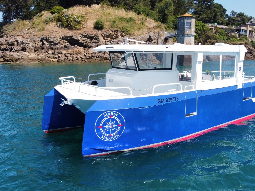 Blue and white catamaran on water near rocky coastline.