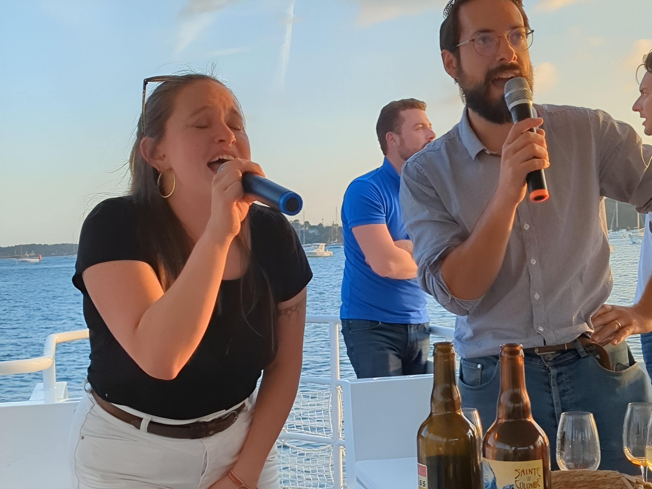 Two people singing on a boat, holding microphones, with bottles on the table.