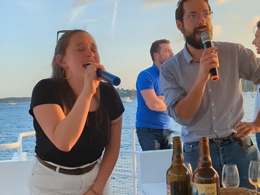 Two people singing on a boat, holding microphones, with bottles on the table.
