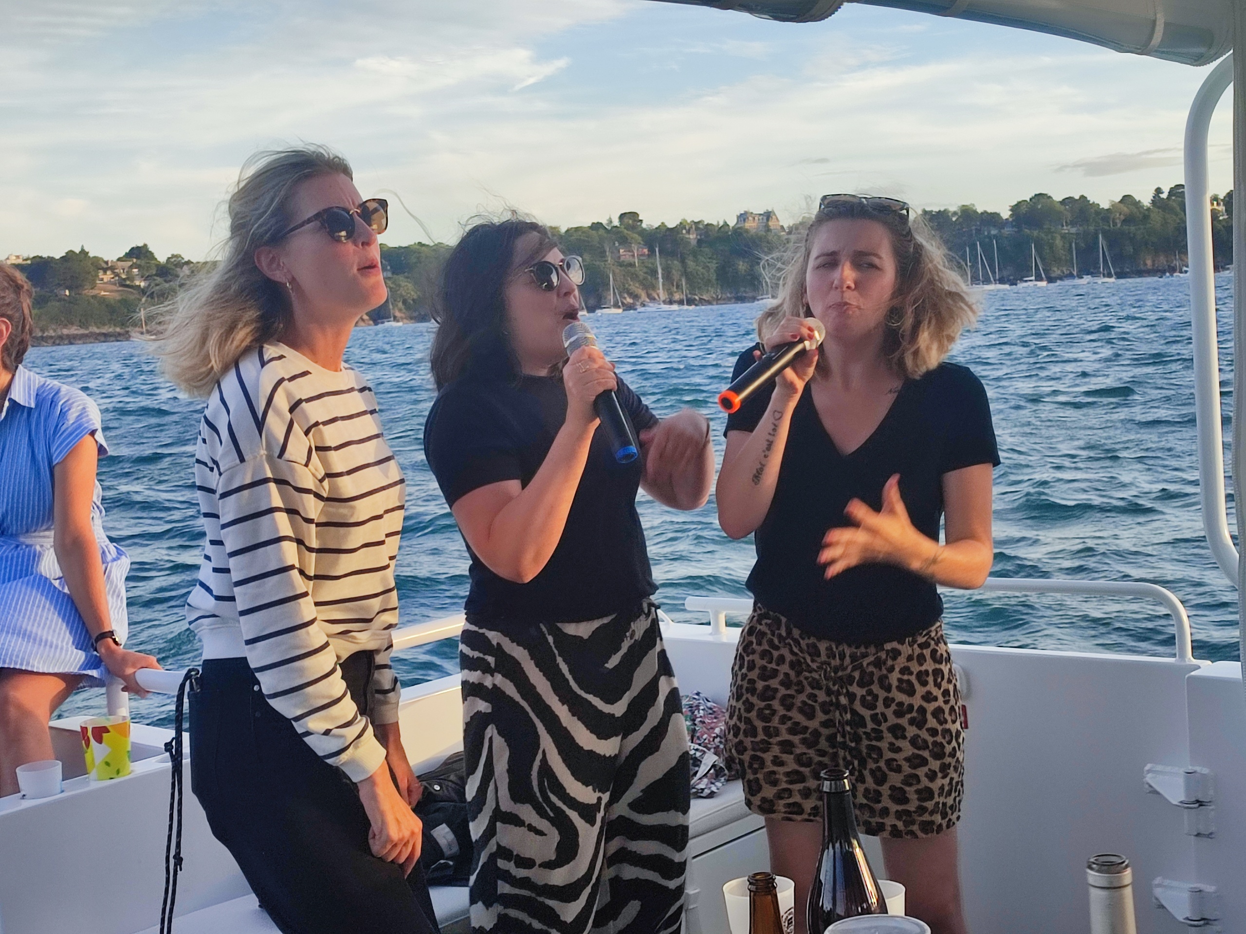 Three people singing on a boat with microphones, ocean in the background.