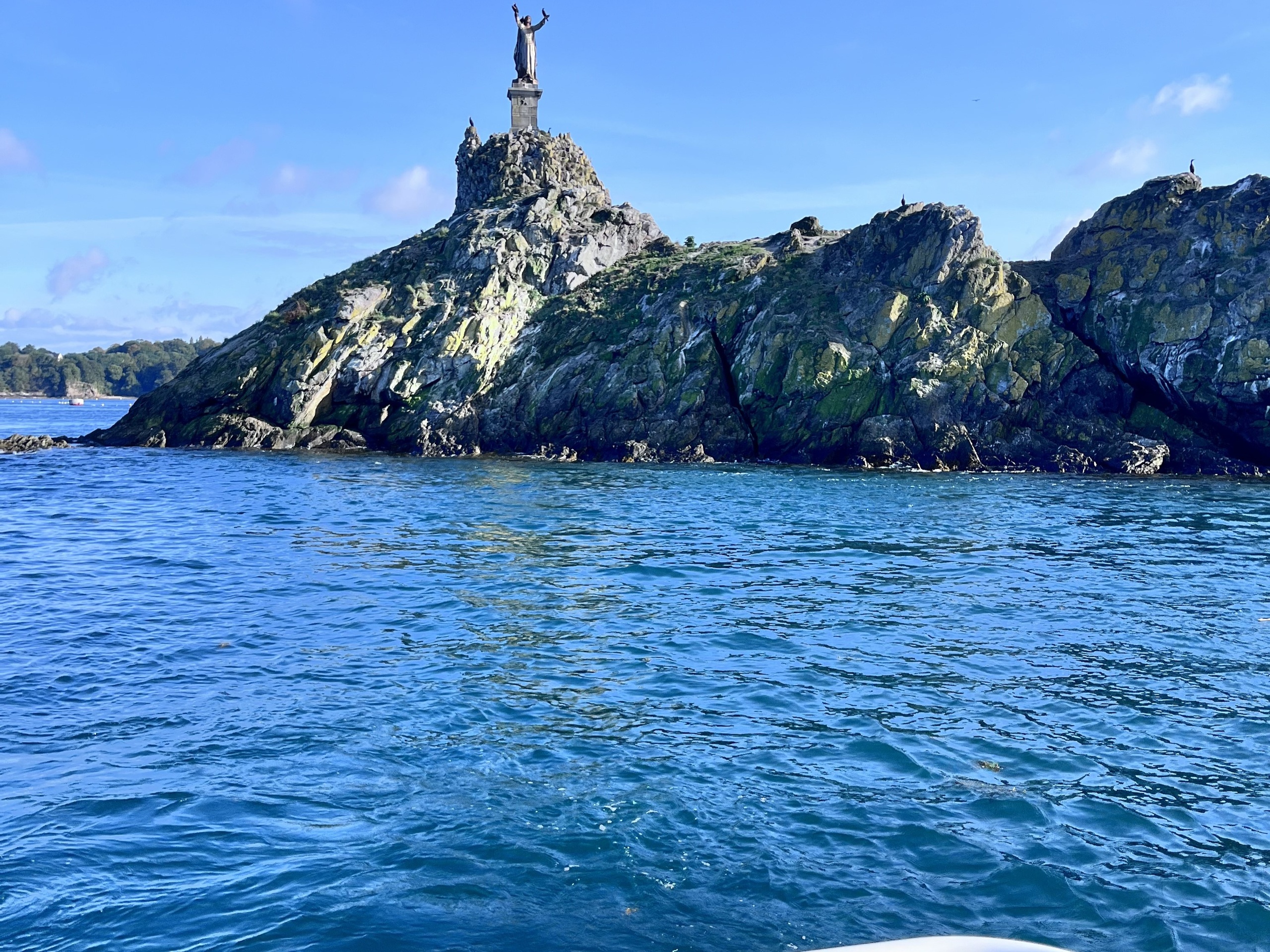 Statue on rocky island surrounded by blue water and clear sky.